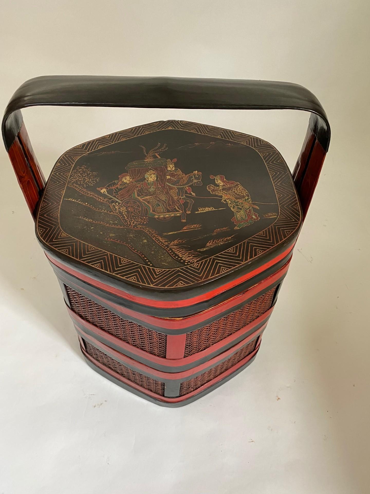 Un antico cesto orientale a tre livelli in legno laccato rosso e nero di forma esagonale, finemente decorato con motivi di cineserie dipinti a mano. La superficie presenta intricate scene dorate e policrome di figure, padiglioni e paesaggi naturali,