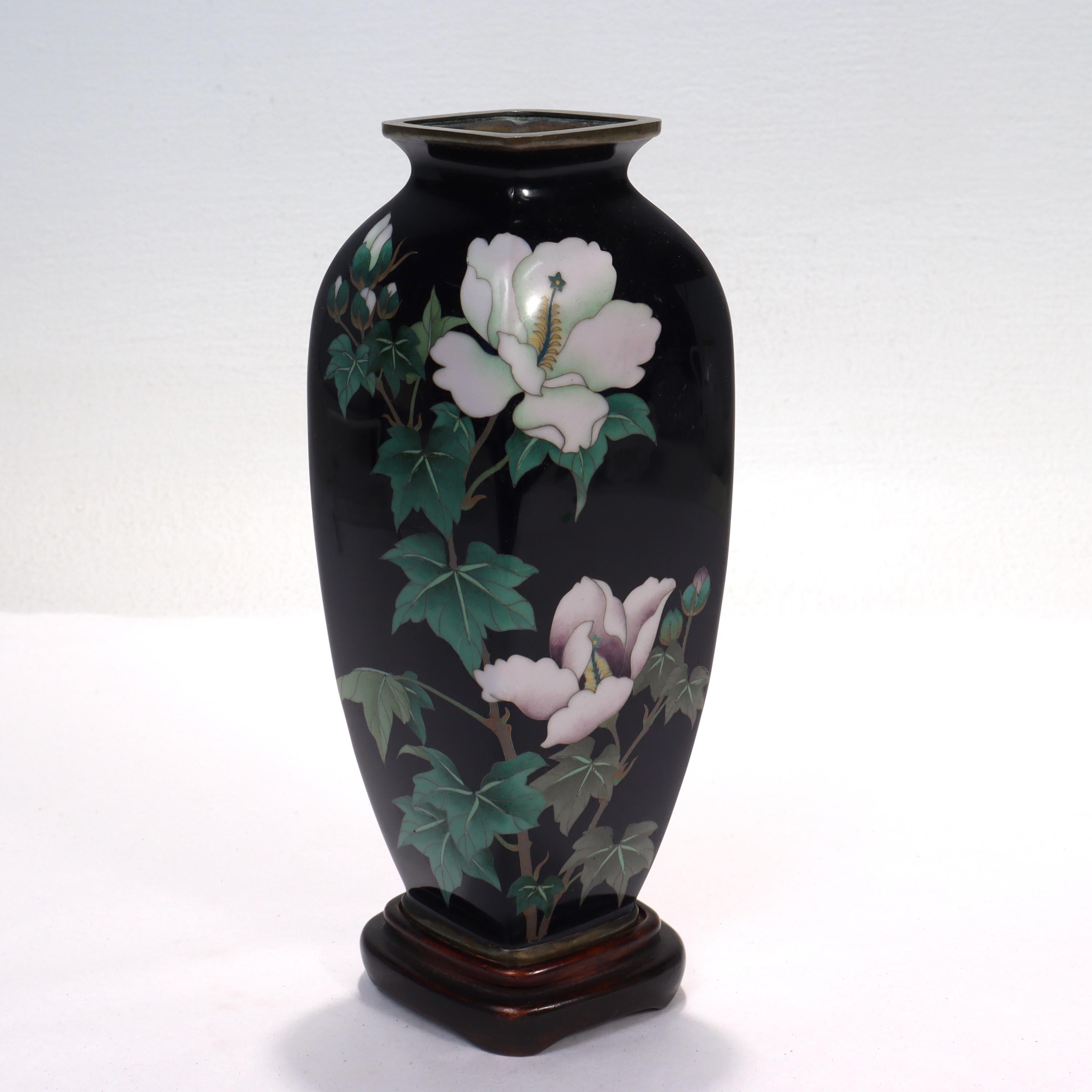 A fine Japanese cloisonne enamel vase.

Decorated with white (stylized morning glory) flowers throughout.

The morning glory - Asagao in Japanese - signifies the bonds of love.

With a trapezoidal opening and base along with conforming wooden