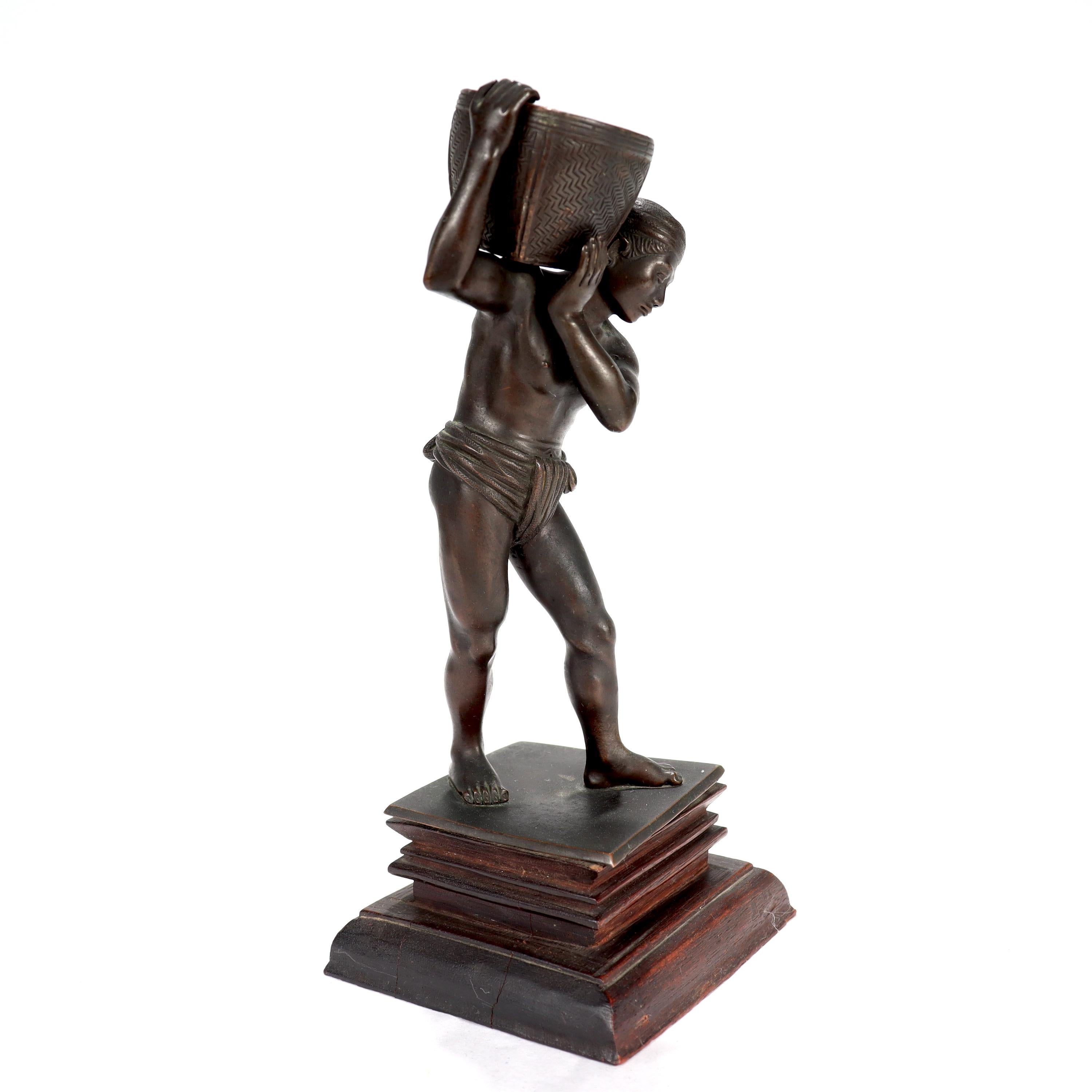 A fine old or antique Pegu bronze sculpture or statue.

In the form of a man in loincloth and headwrap holding a basket on his right shoulder (perhaps a water carrier). 

Secured to a conforming, carved wooden plinth with a bolt.

(The basket