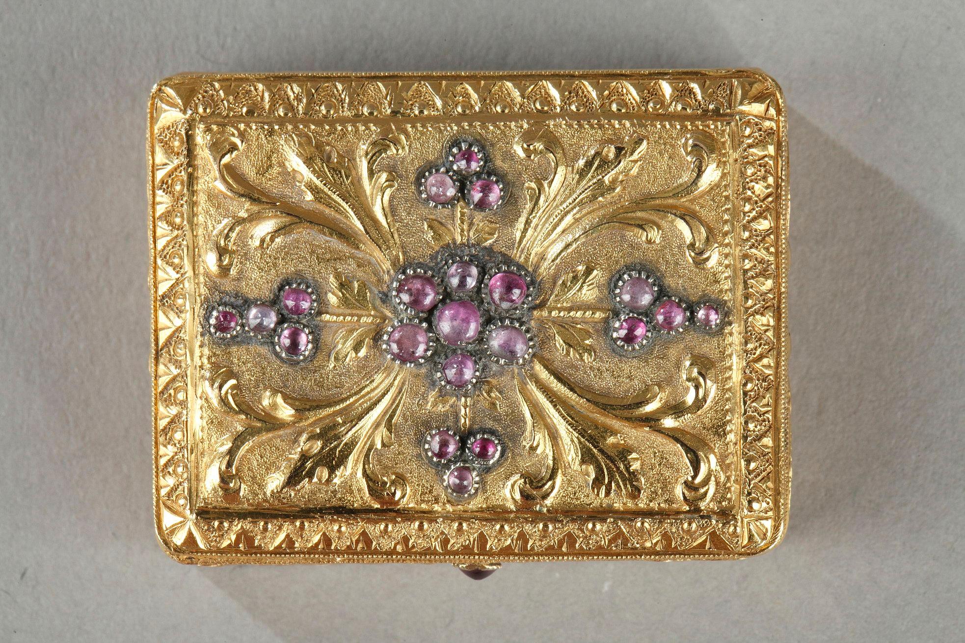 Rechteckige, goldene Vinaigrette mit Scharnierdeckel. Reliefartige Schnörkel heben violette Cabochon-Steine hervor, die in der Mitte zu einer Blume angeordnet sind. Ein geometrischer Fries umrahmt die Komposition. Der Korpus der Dose ist mit