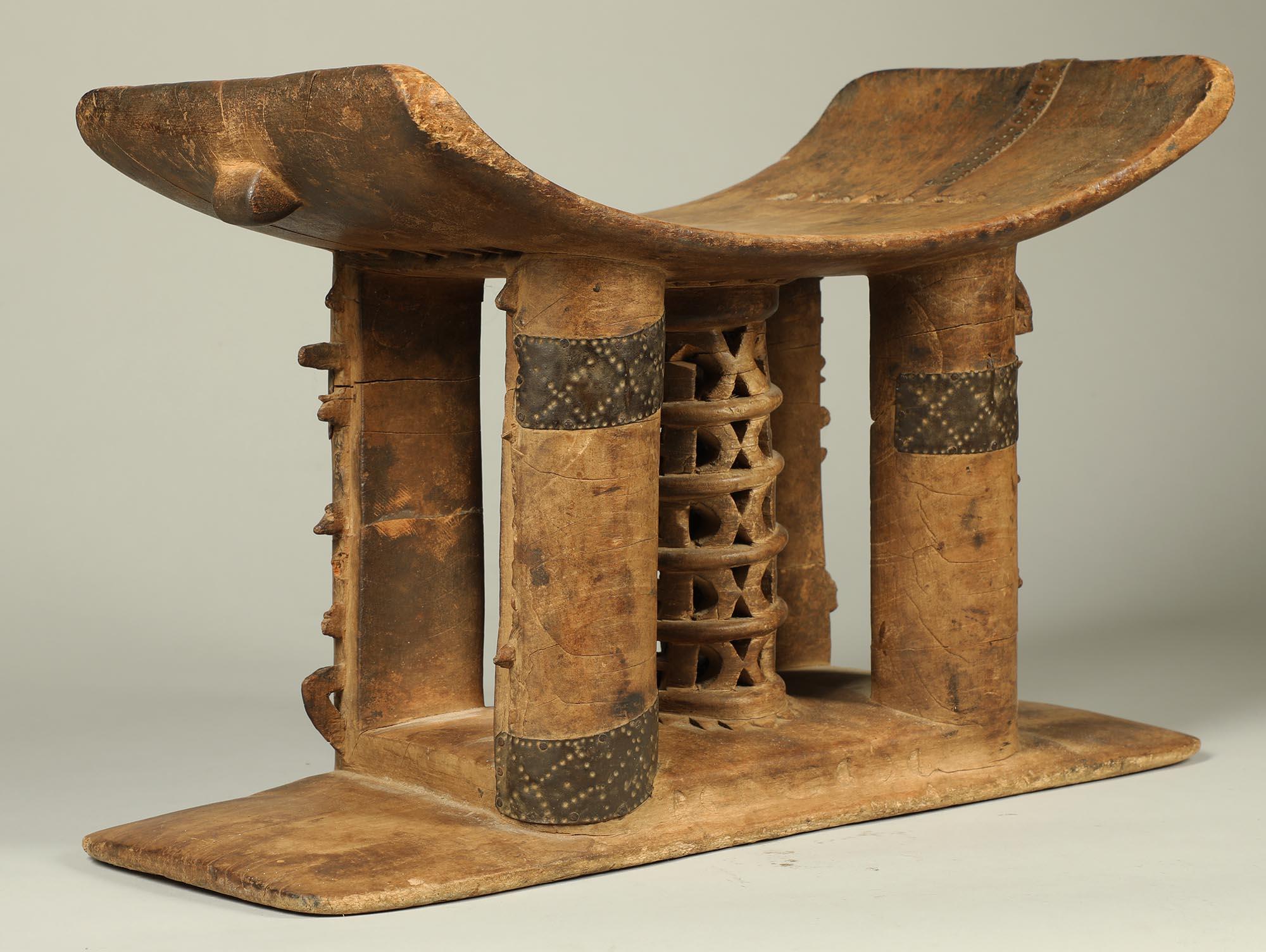 The stool was loved by its owner, repaired in several places with decorated metal straps. It has an openwork, tiered lattice central post and four support posts. It has the older style tiered square cut out and wide cross hollow on the underside