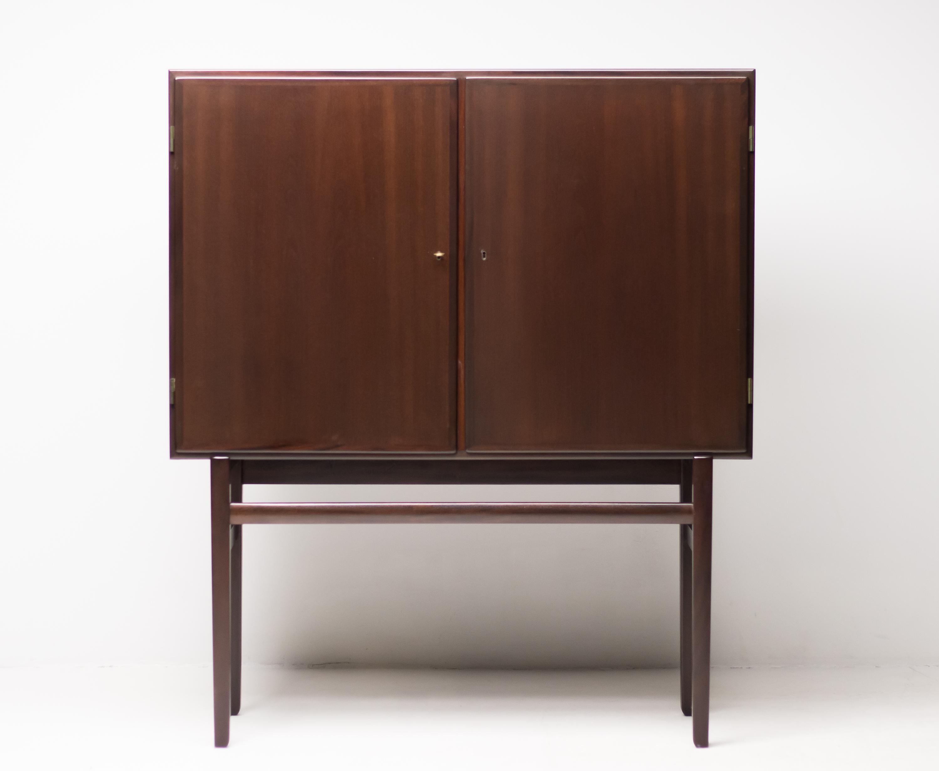 Elegant upright 1950s cabinet in beautifully grained mahogany.
Two doors conceal four interior shelves and one drawer over four legs with high stretchers.
Includes the original brass key for the door locks.
Ole Wanscher and Danish Makers tags on