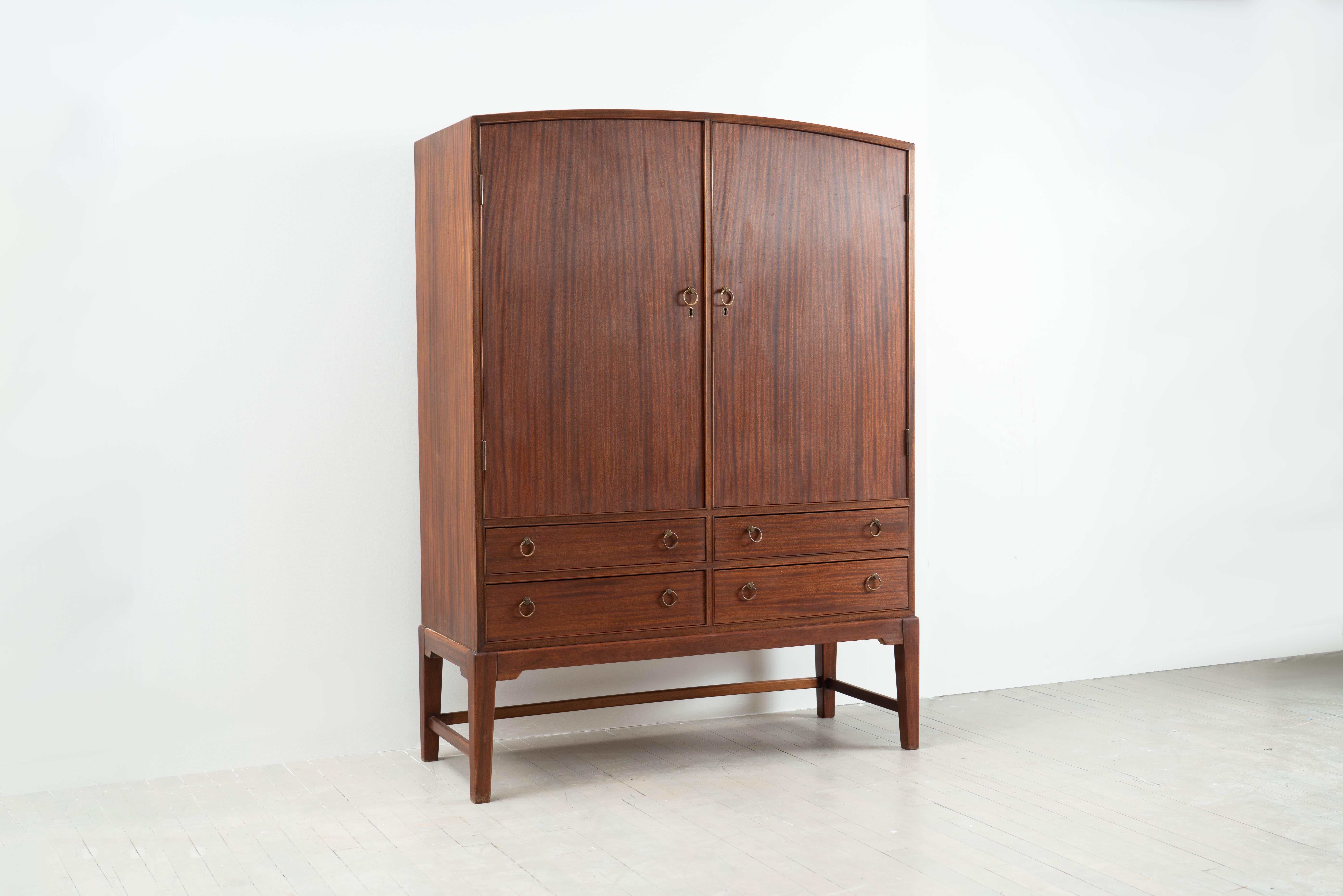 This beautiful arched top mahogany cabinet by Ole Wanscher retains its original oiled finish and brass hardware. Two keyed doors open to reveal adjustable shelves and four lower drawers offer additional storage.