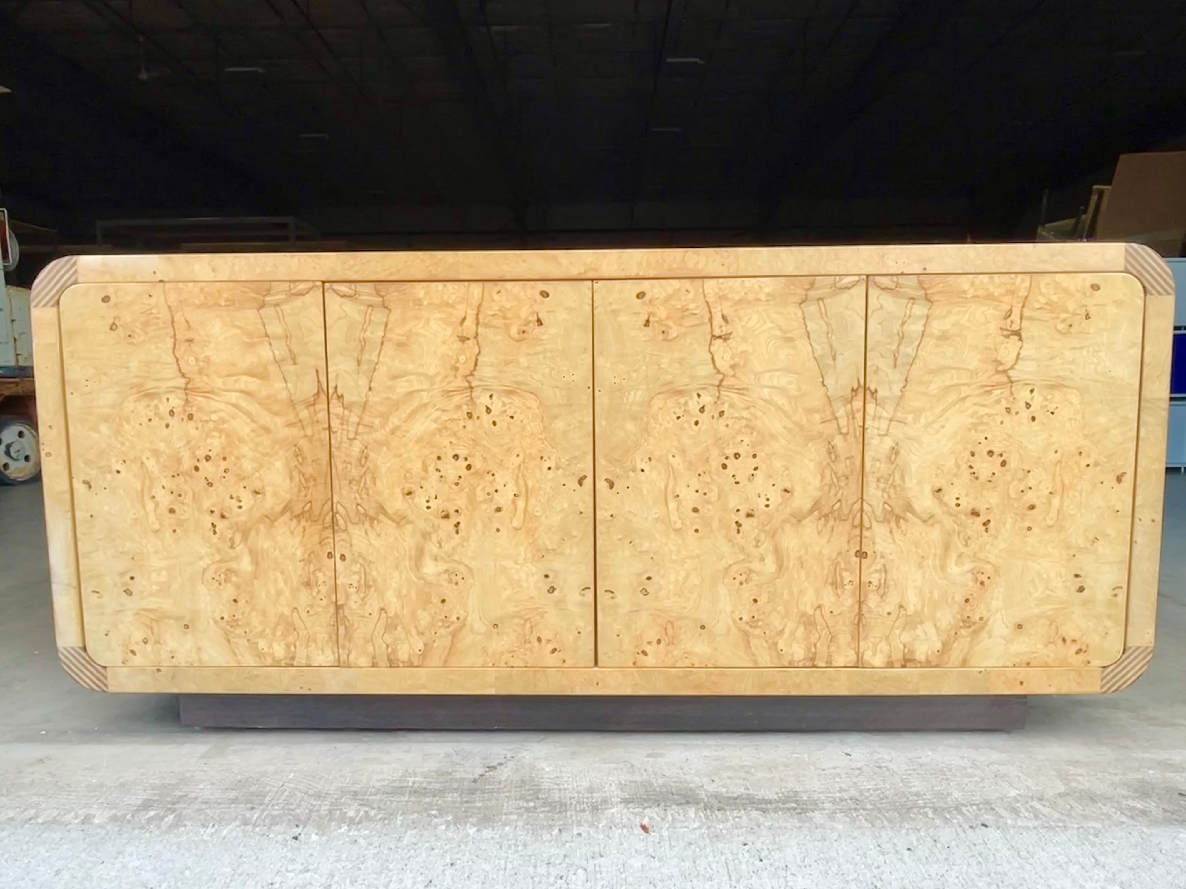 Henredon scene two credenza sideboard buffet cabinet in olive ash burl bookmatched veneer with zebra wood radiused corners and four flush doors with spring clip closures. Circa 1985.
Divided into two compartments each with a single drawer and shelf.