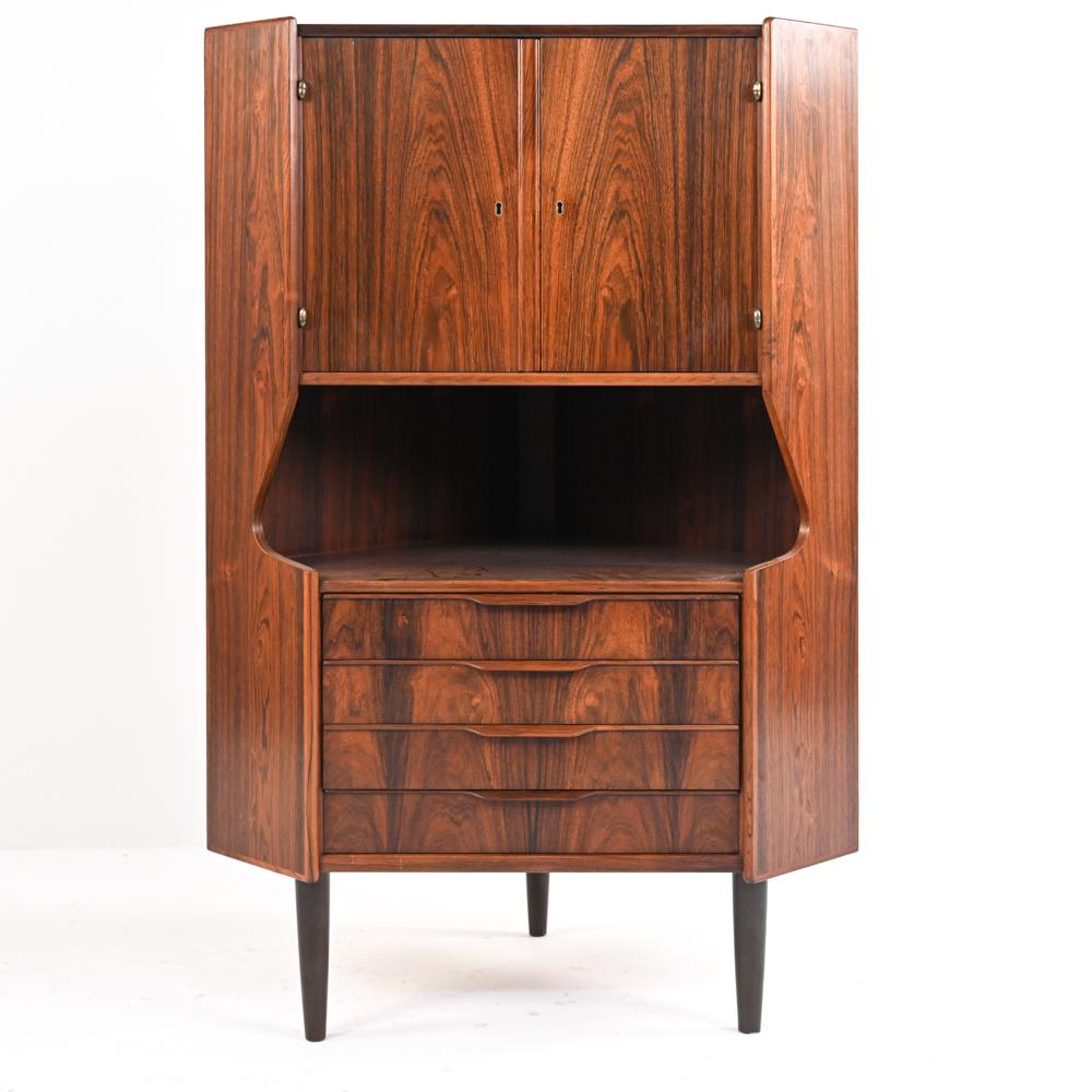 A well-proportioned and multifunctional corner cabinet in dramatic Brazillian rosewood, with dovetailed mahogany and pine plywood drawers, carved wing handles, and a pinstriped mirrored interior. Designed and produced by Omann Junn, c. 1960's. Key