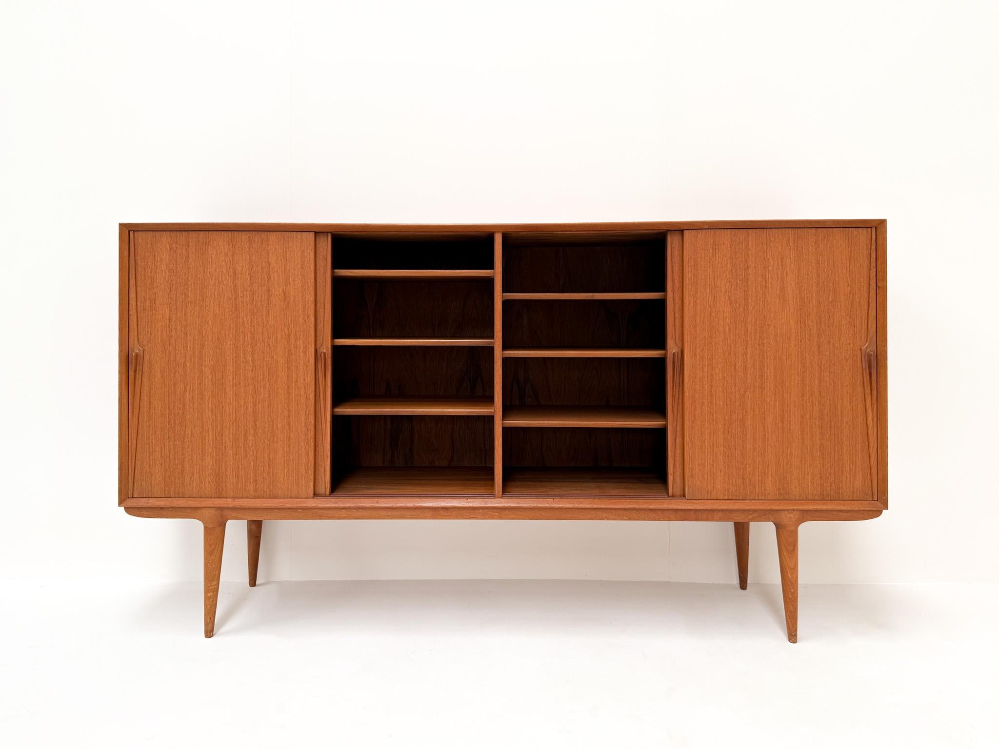 This elegant mid-century sideboard was designed by Gunni Omann for Omann Jun in Denmark during the 1960s. It is made from teak wood and features four sliding doors with sculpted solid wood handles. Inside, it offers a mix of adjustable shelves and