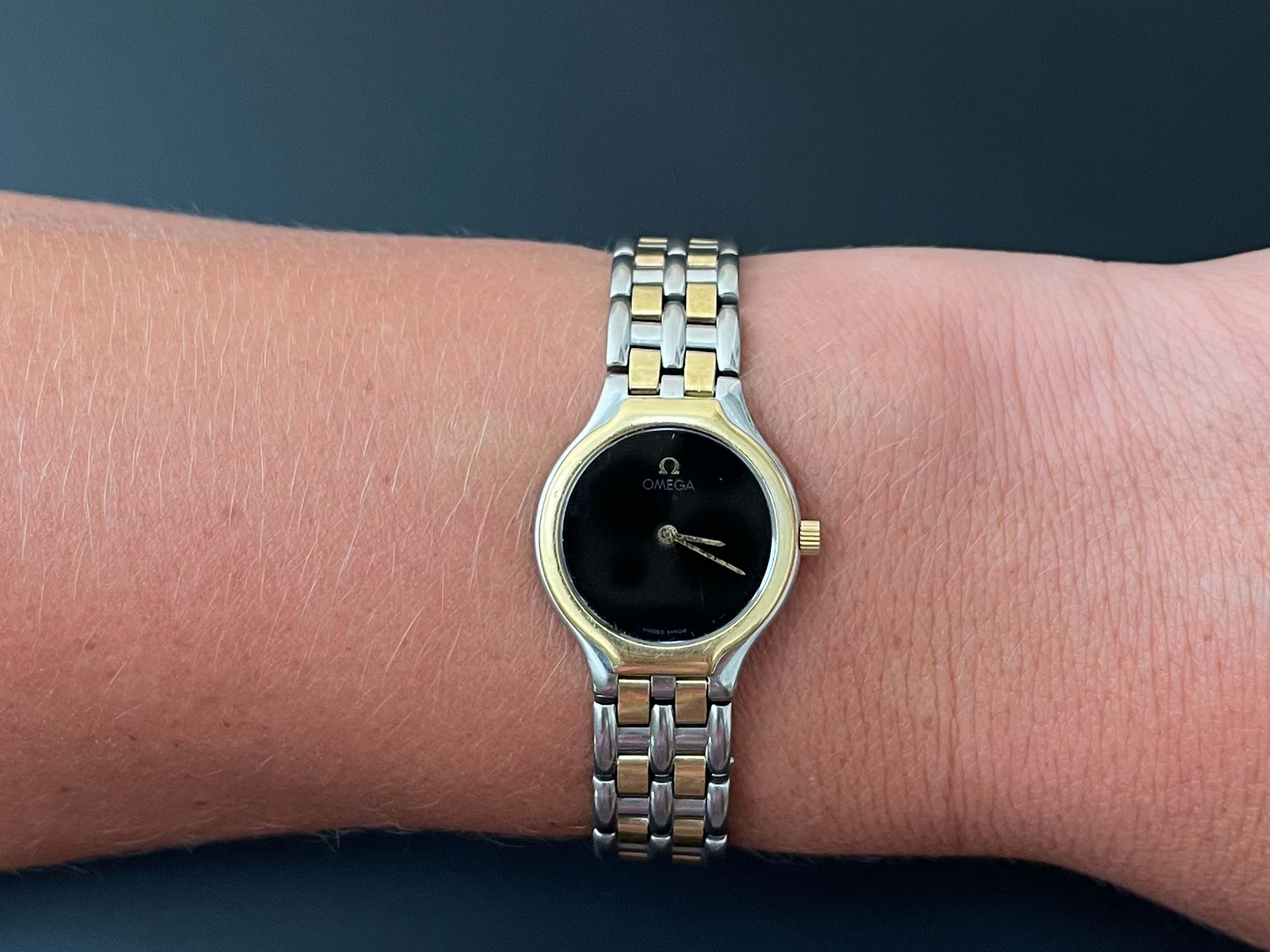 Omega 18K und Stahl Damen DeVille Uhr. Diese klassische Armbanduhr ist charismatisch und fesselnd. Sie hat ein rundes 23-mm-Gehäuse und ein schwarzes Quarzzifferblatt mit Stunden- und Minutenzeiger. Die Uhr hat eine Lünette und Krone aus 18 Karat