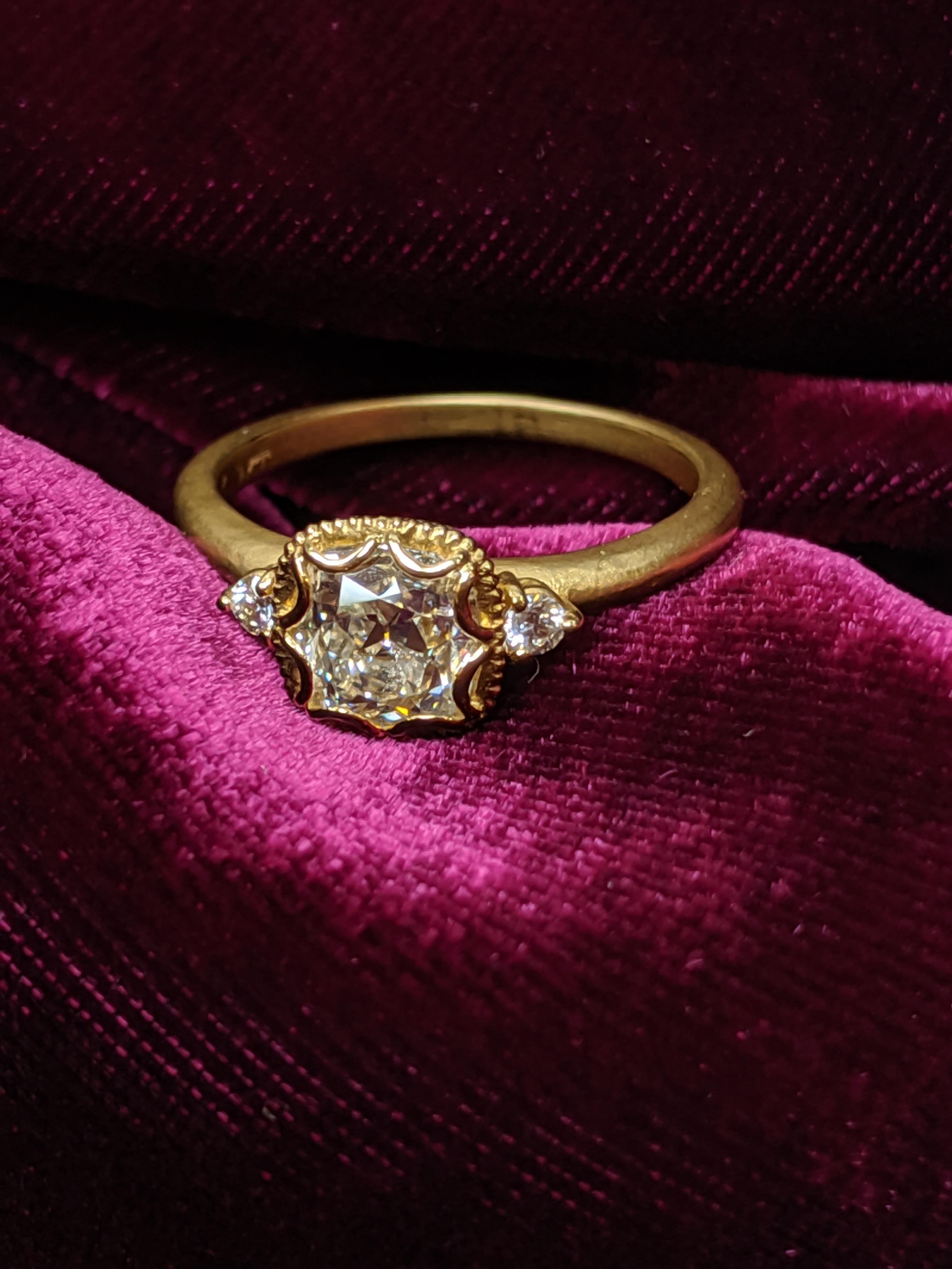 As if out of yesteryear a sweet Antique Cut Cushion Diamond ring in an 18 karat Yellow gold.  Center diamond is 1.07 carats with a GIA Report J VS1.  On either side are single smaller accent round brilliant diamonds.  This ring mounting is