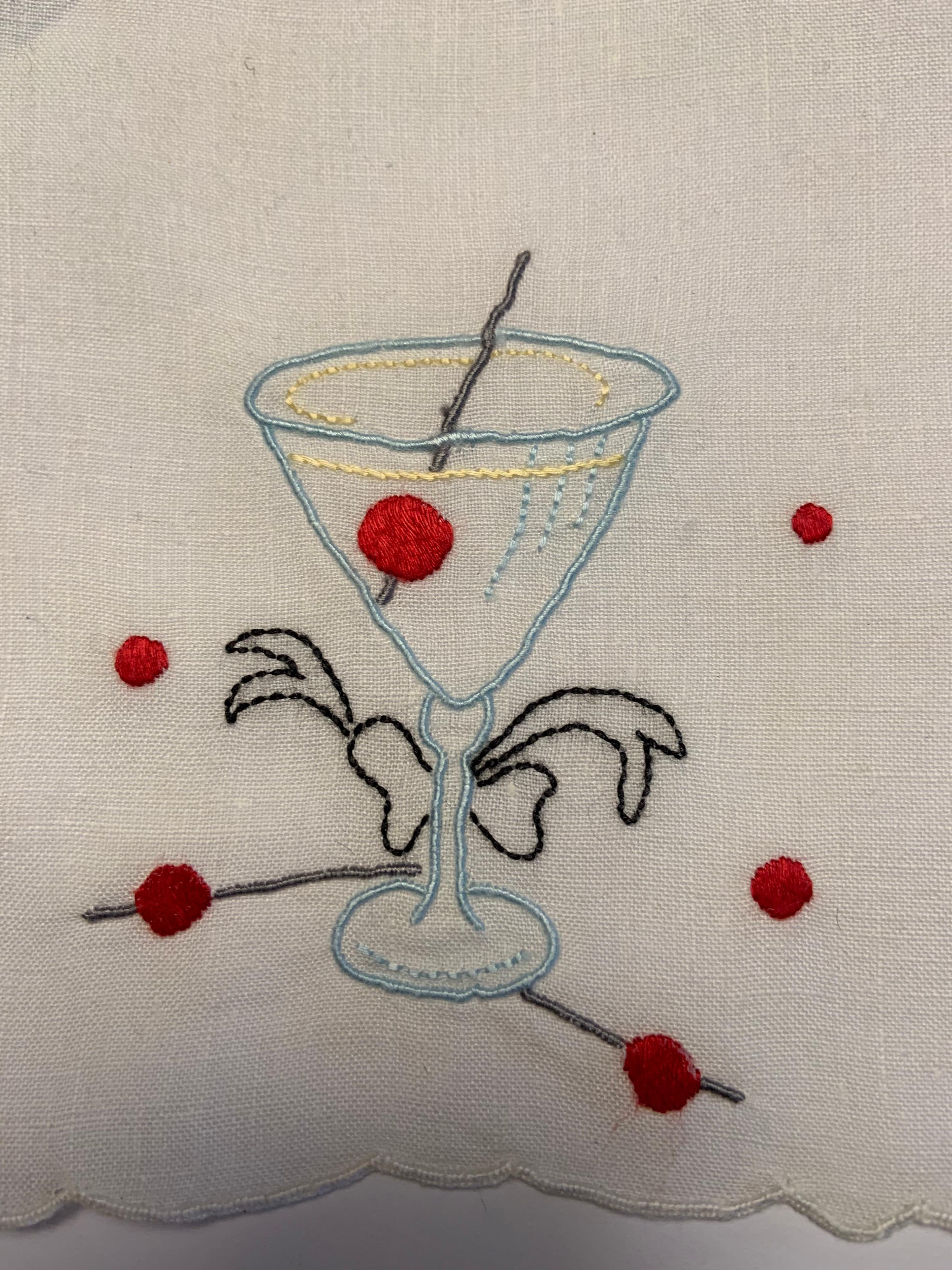 A sleek stemmed Manhattan cocktail glass is embroidered in blue with a red cherry and grey stirrer inside on this set of twelve white cocktail napkins. They have embroidered red dots adding to the festive look and they are all linen with scalloped