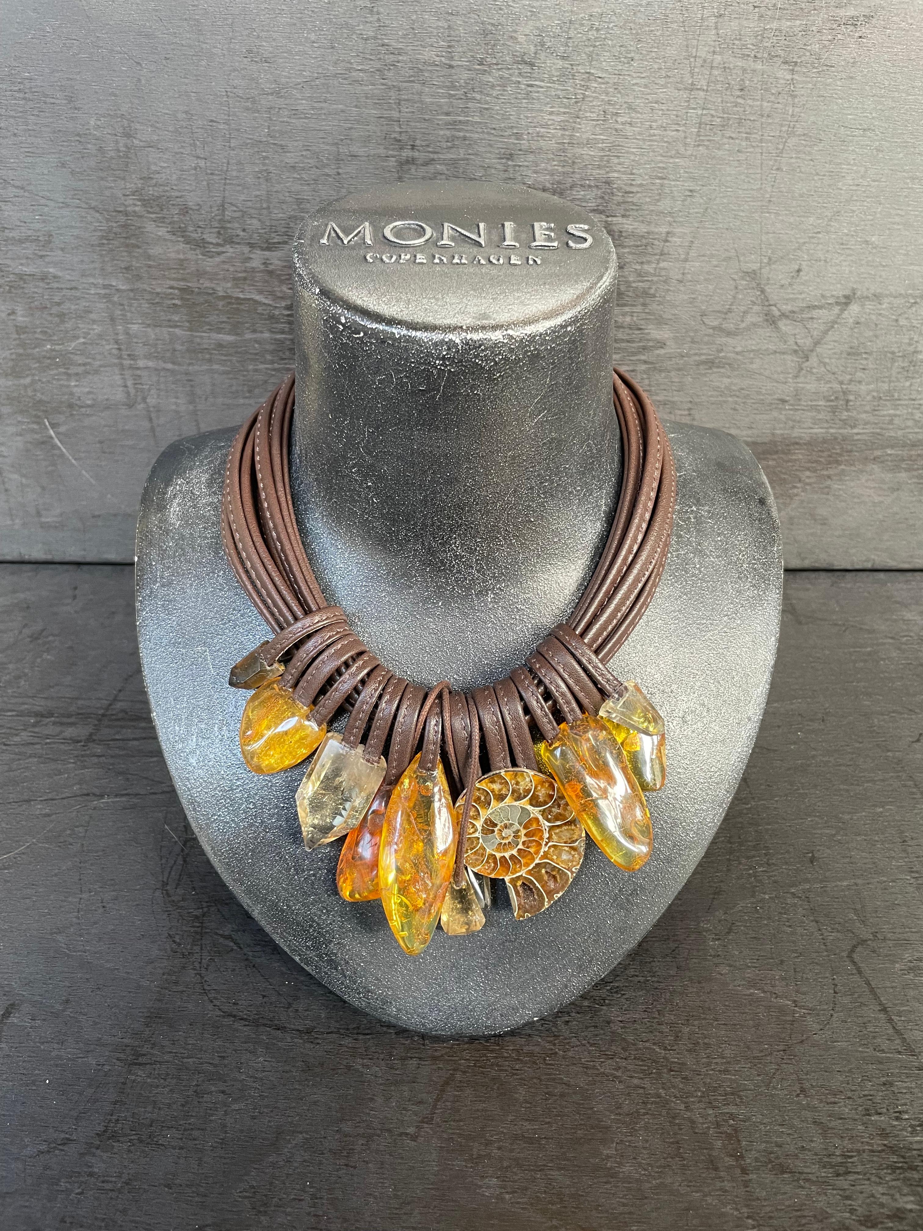 Collier unique de la marque de bijoux danoise Monies.
Fabriqué en ambre, ammonites, cristal et cuir avec un fermoir en cuir.

Fabriqué à la main dans l'atelier de Monies à Copenhague.