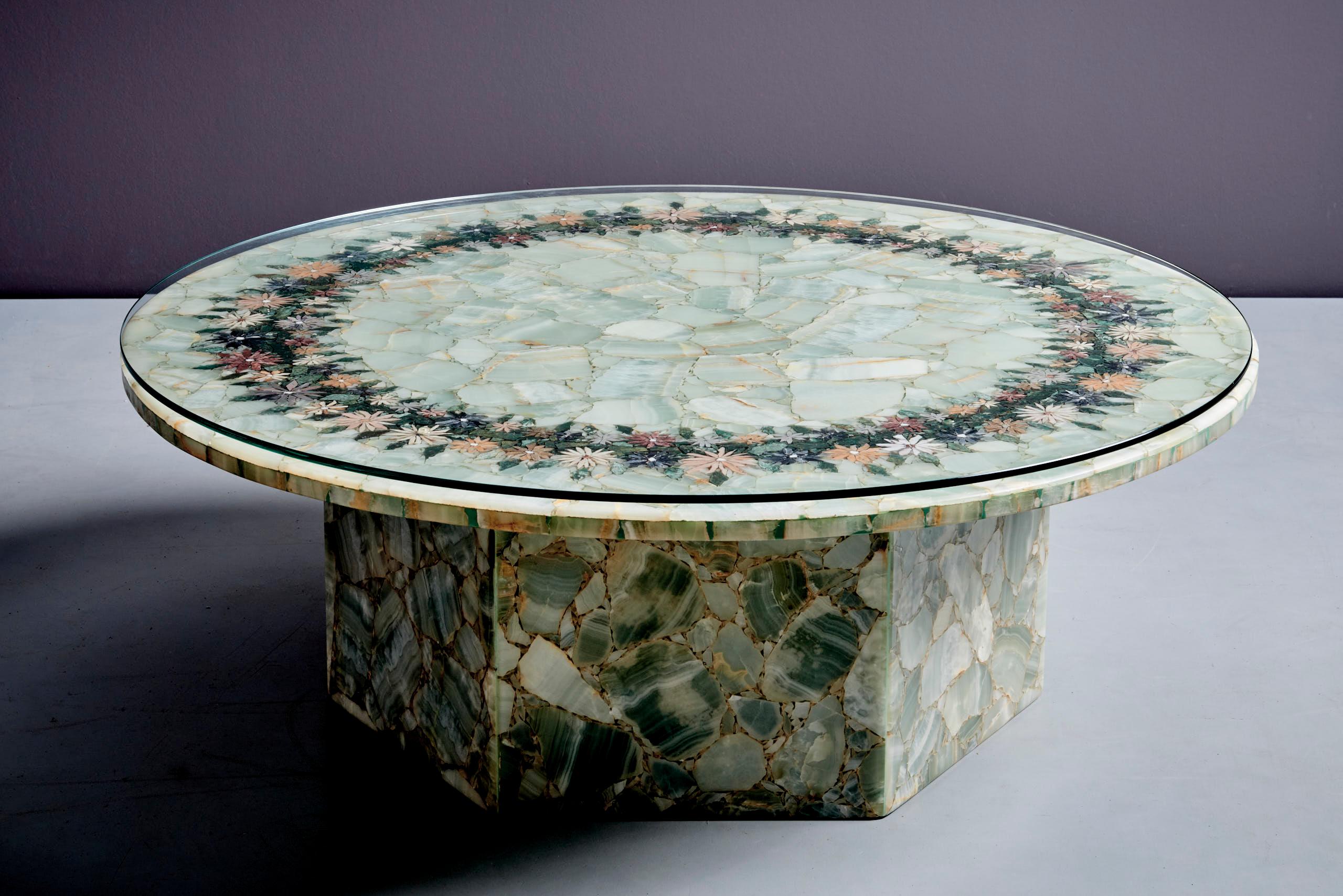 Esta elegante mesa redonda está hecha de ónice verde con un impresionante mosaico floral de piedra en su superficie. La intrincada incrustación de piedras de colores forma un delicado anillo de flores, mostrando un arte extraordinario. Esta mesa