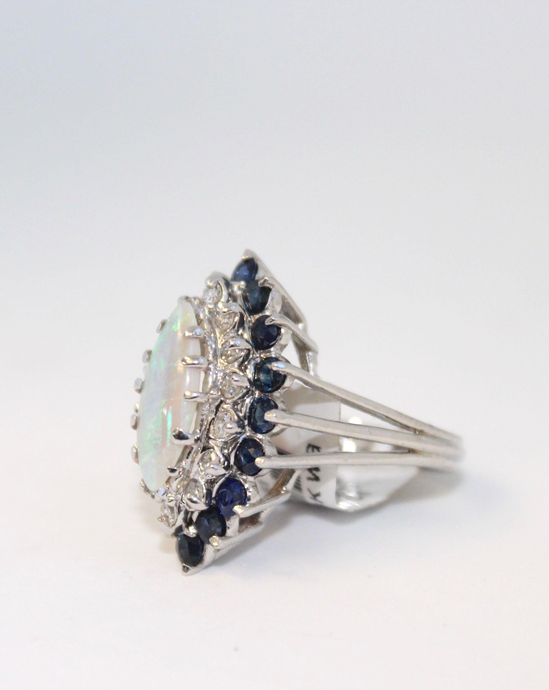 This stunning ring is set in 14K White Gold with One (1) color-filled opal (dimensions 15 x 8 mm). It contains fourteen (14) Round Single Cut Diamonds Weighing 0.21ctw and Sixteen (16) Round Blue Sapphires weighing 1.60ctw. 

It is currently sized
