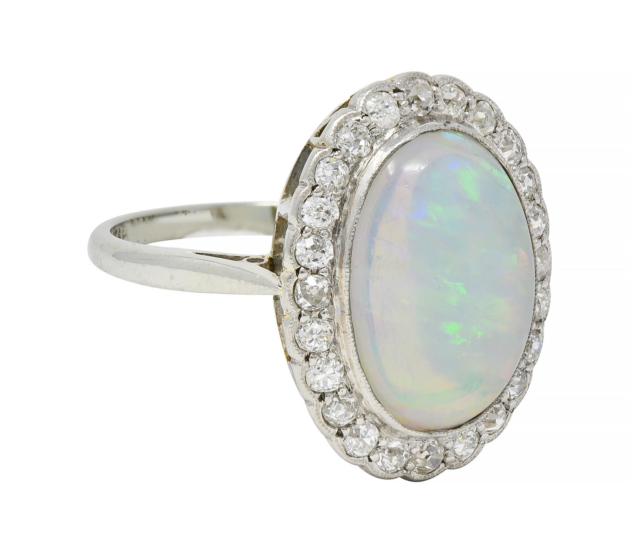 Centering an oval-shaped jelly opal cabochon measuring 10.5 x 13.0 mm bezel set in platinum-topped gold
Transparent white in body color with spectral play-of-color 
Featuring a halo surround of old European cut diamonds 
Weighing approximately 0.72