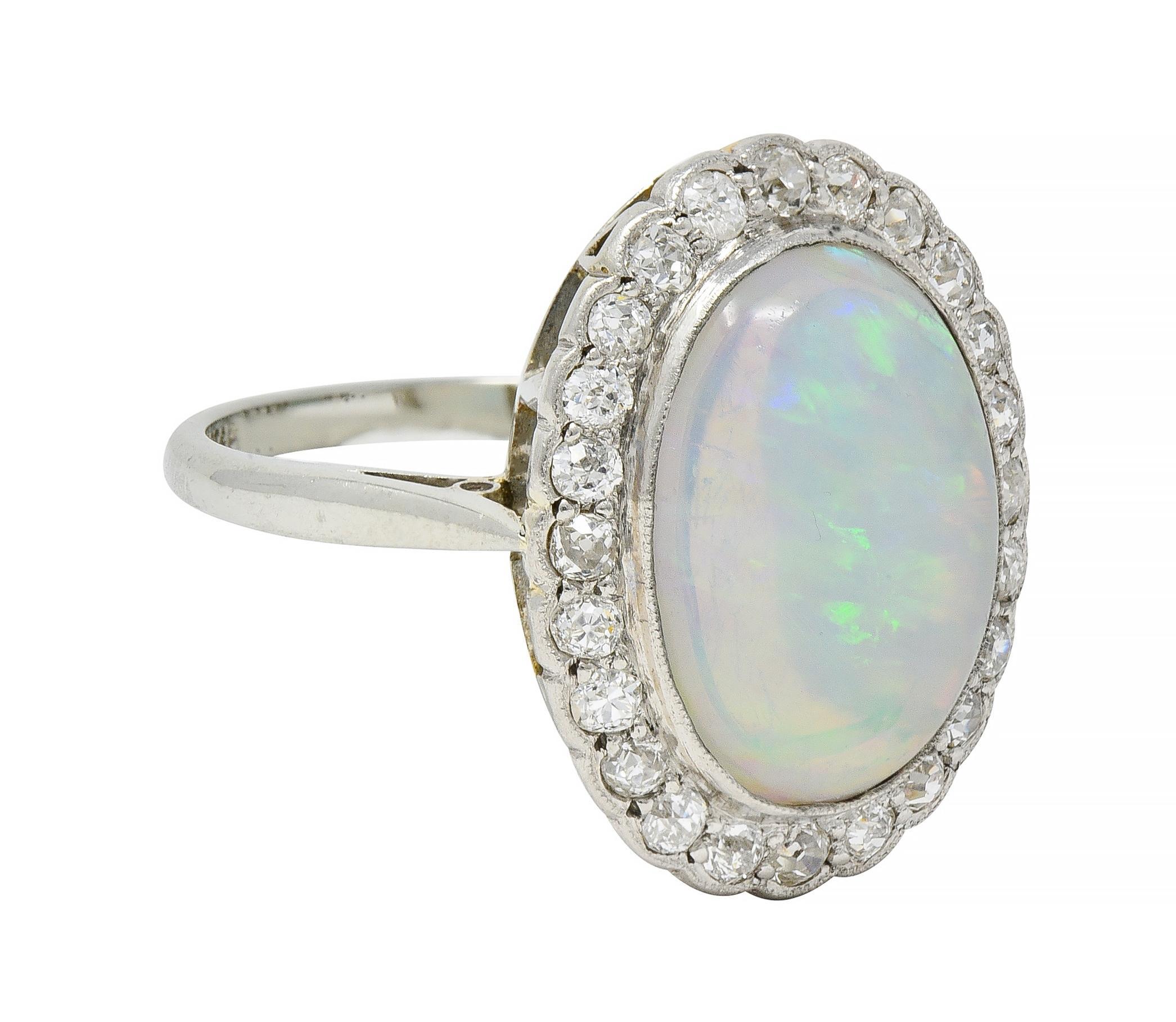 Centra un cabujón de ópalo gelatinoso de forma ovalada de 10,5 x 13,0 mm engastado en bisel en oro con baño de platino
Blanco transparente en el color del cuerpo con juego espectral de colores 
Con un halo rodeado de diamantes de talla europea