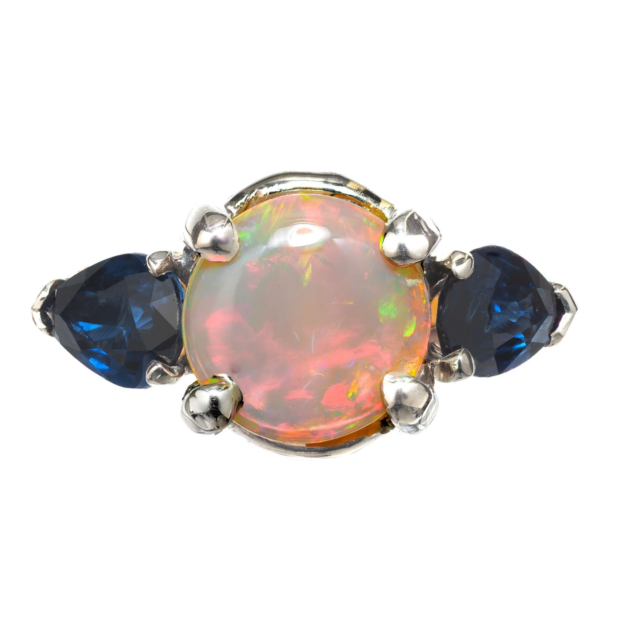 Orange, red, blue and green opal engagement ring with two side sapphires. The center stone is set in a 14 white gold basket and 14k yellow gold three-stone setting. 

1 round opal approx. total weight 1.00cts. Opal is 7.4mm across
2 pear shape