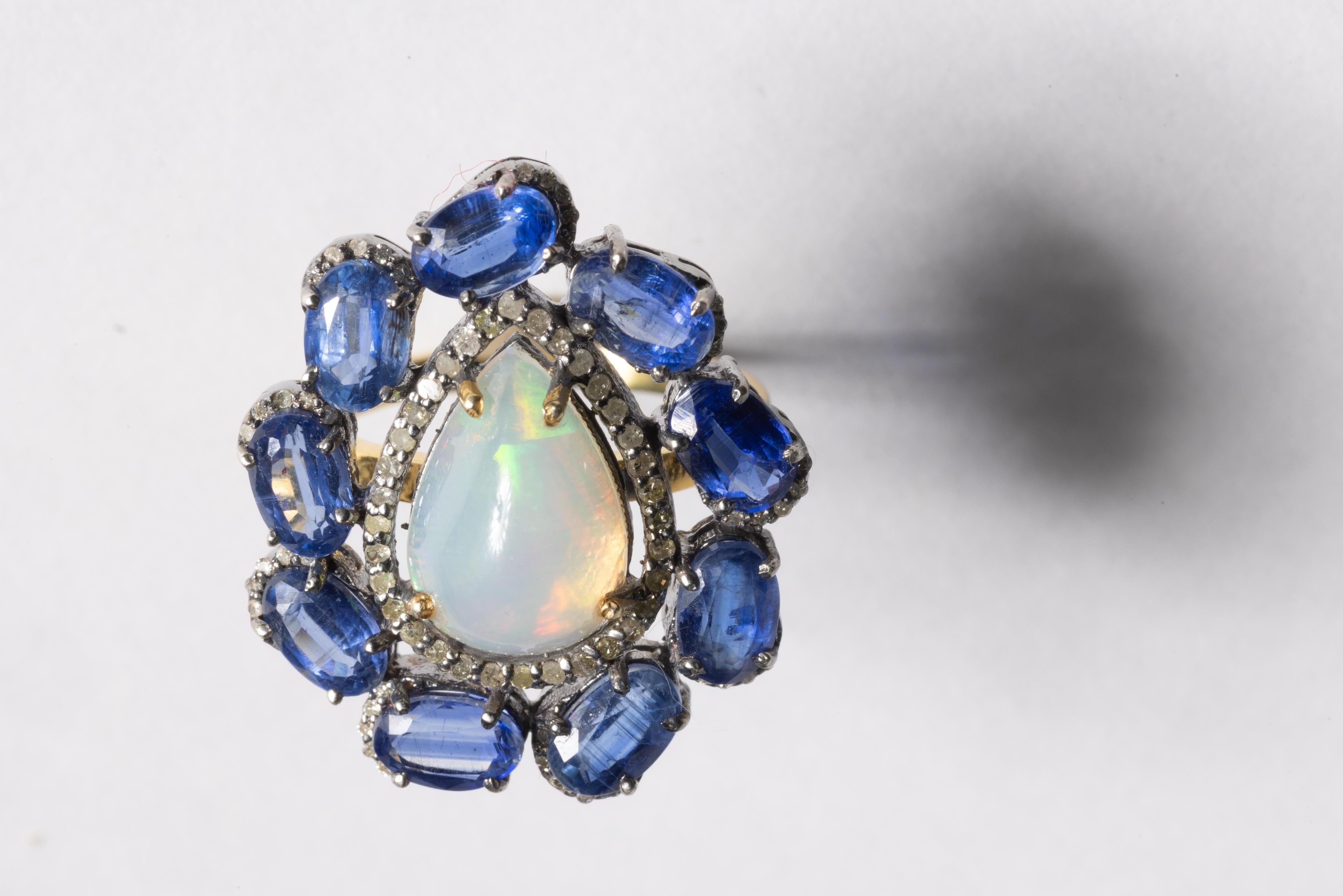 An amazing cocktail ring featuring a large, pear-shaped opal at the center in 2.10 carats.  It is bordered in round, brillilant cut diamonds and then bordered again with faceted, oval kyanite gemstones with an apron of diamonds around each