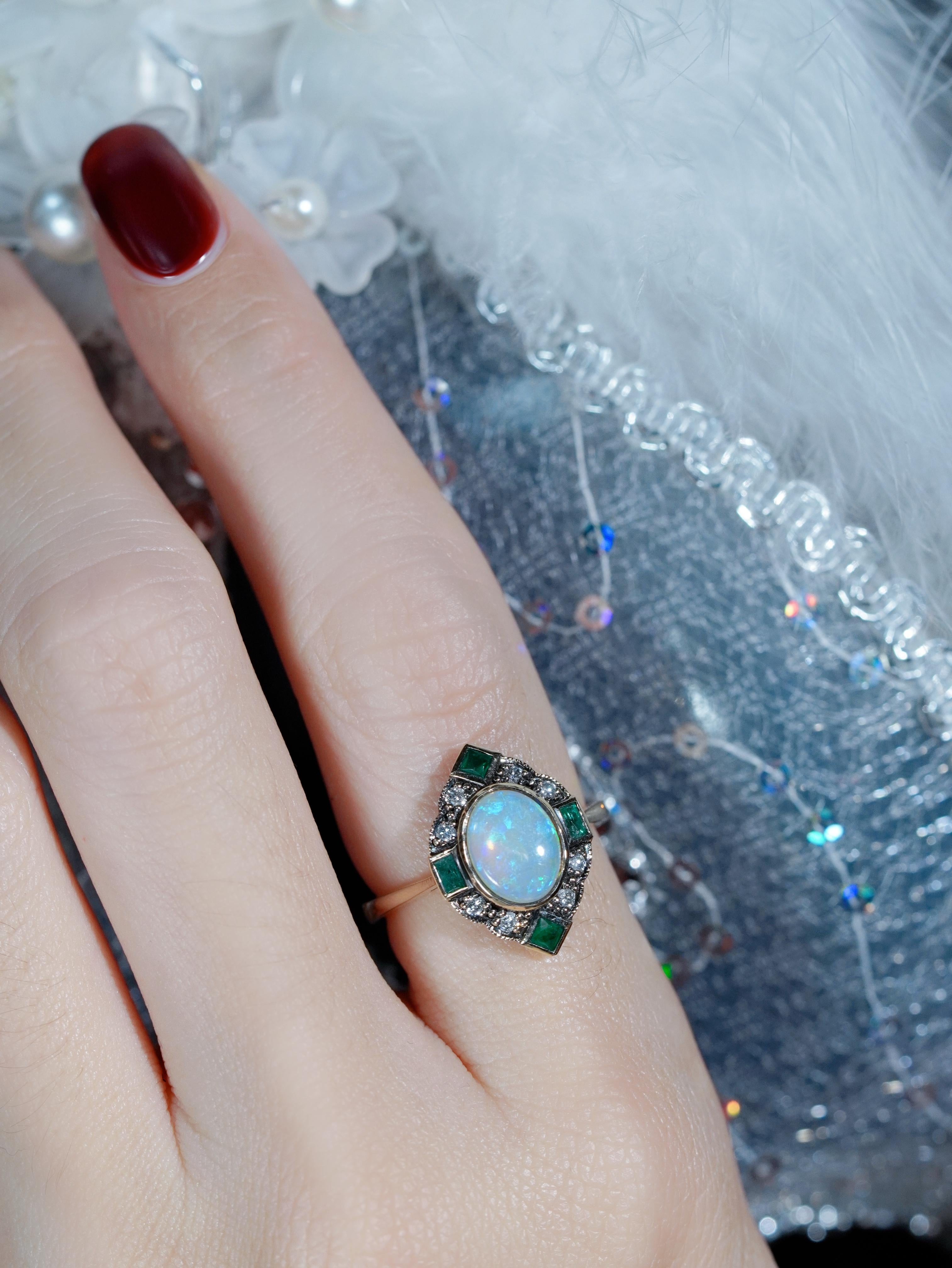 Cette bague halo Art Deco, qui brille d'une allure vintage, célèbre la couleur et la lumière. Une opale incandescente repose dans une lunette lisse, entourée d'un halo étincelant de diamants. Quatre émeraudes rayonnantes, placées en haut, en bas, à