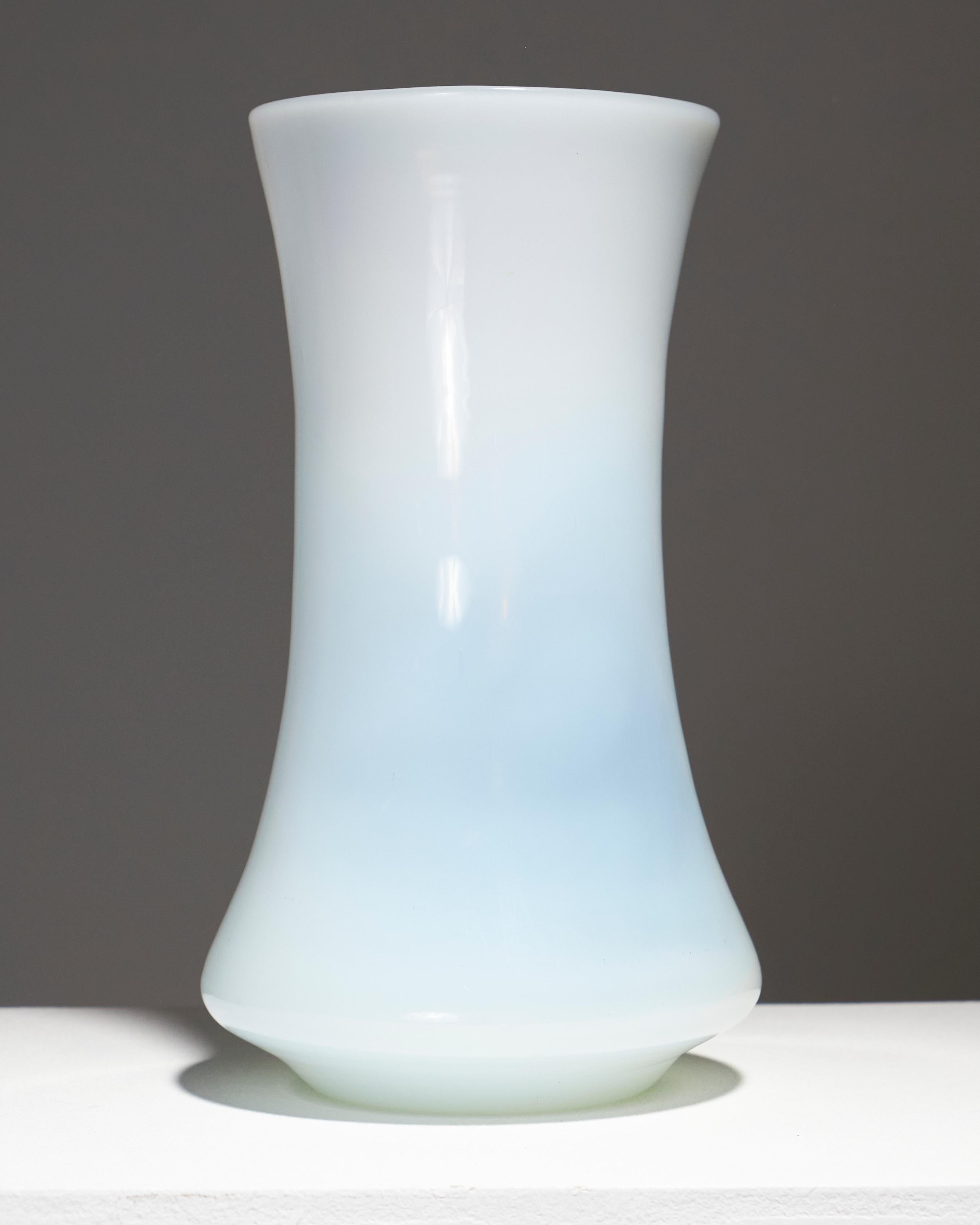Vaso in vetro bianco opalescente degli anni '60, di forma cilindrica, leggermente affusolato al centro e svasato verso il collo. Il suo design elegante e senza tempo evidenzia la morbidezza delle sue curve e la sottigliezza della sua trasparenza. In