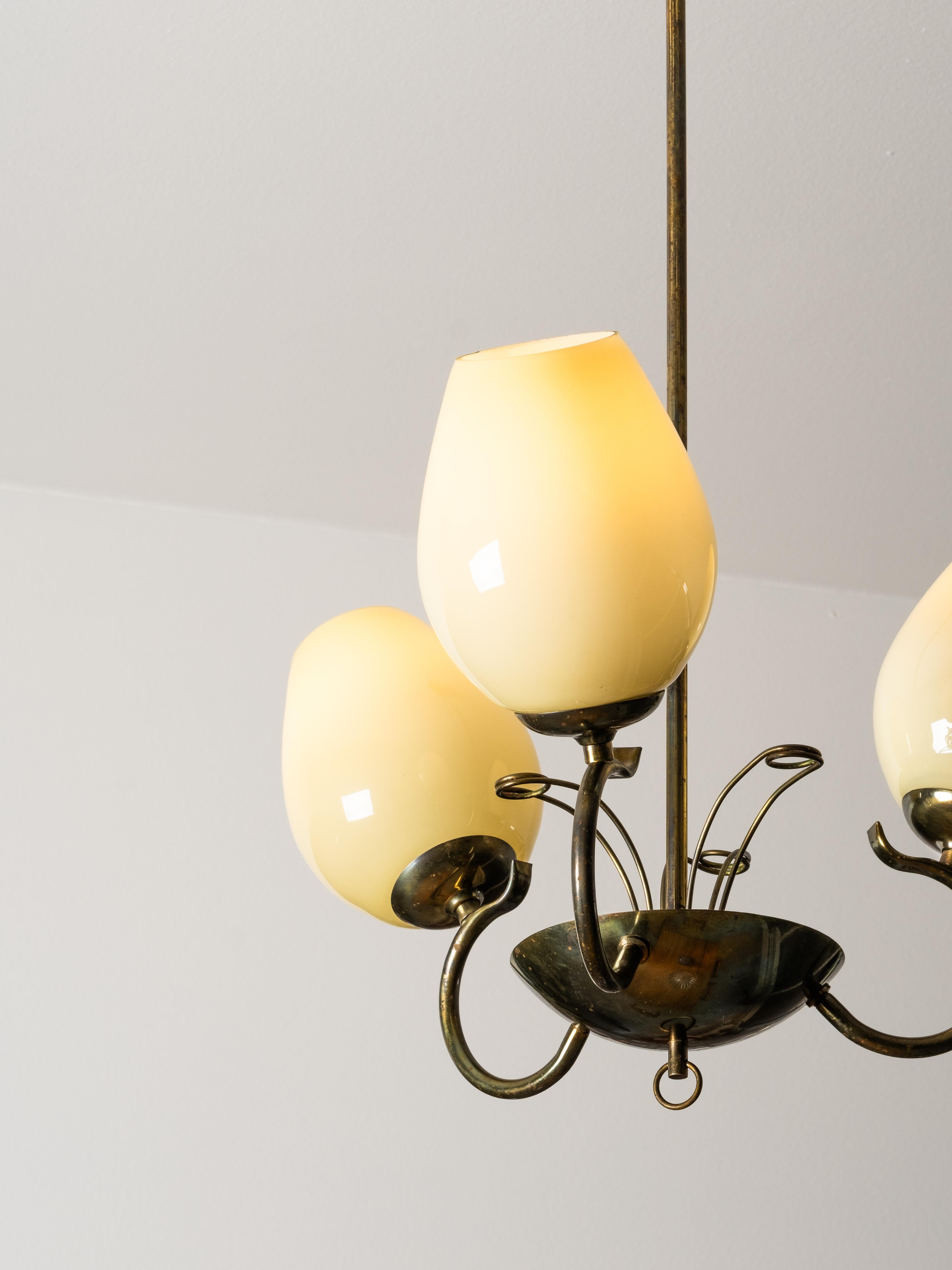 Un lampadario finlandese della metà del secolo scorso, realizzato negli anni '50 da Sievä. La lampada presenta tre paralumi in vetro opalino bianco crema soffiato a mano, ciascuno montato su bracci in ottone graziosamente curvati. La struttura in
