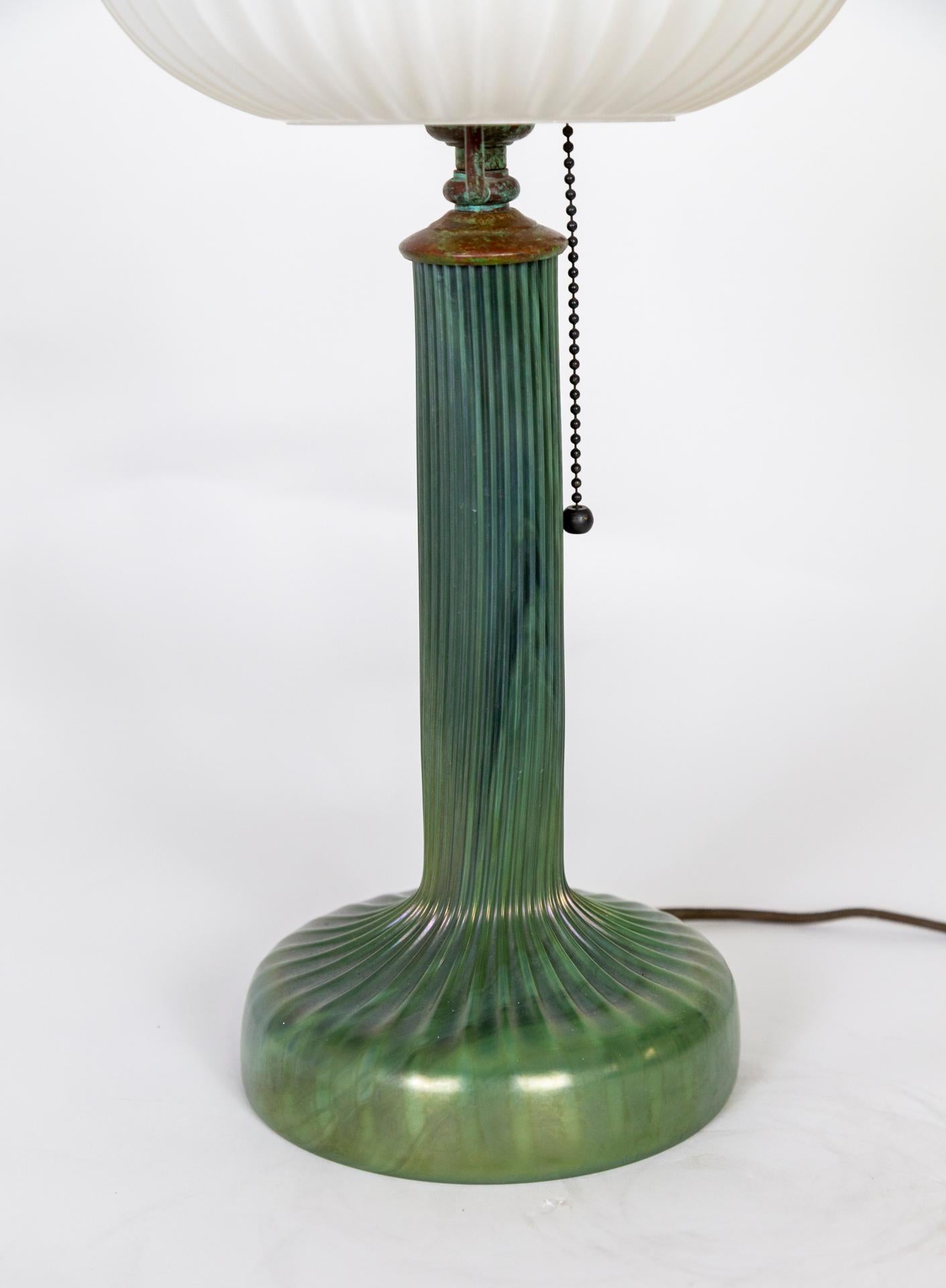 A unique, finely crafted lamp with an opaque, variegated green glass body with reeded details and metal accents with multicolored patina. The matte, milk glass shade is an elliptical, urchin shell shape, with ribbed texture. With a pull chain