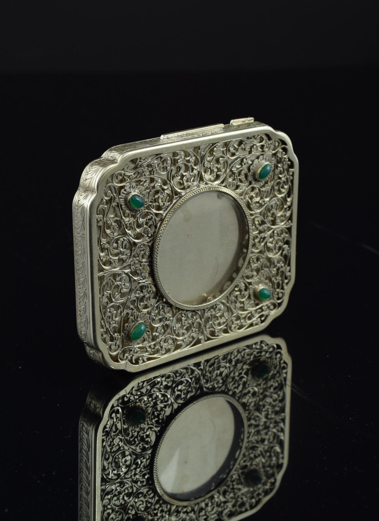 Rectangular silver box decorated with a delicate work of fine curved stems and leaves on the lid, arranged around a circular central space and with four elements flowered with green stones towards the corners. It emphasizes the symmetry of this