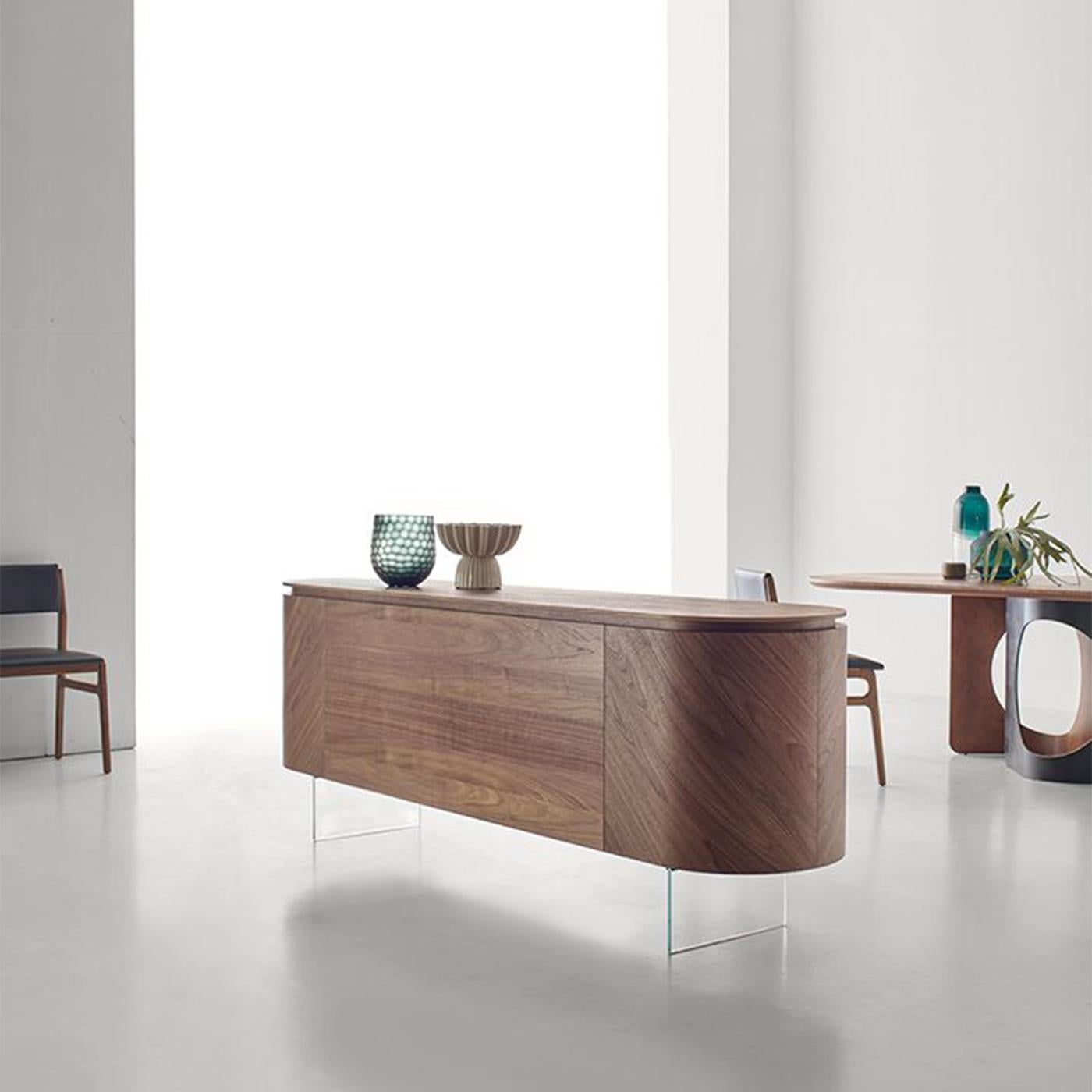 Enveloping curves trace the harmonious silhouette of this exclusive sideboard seemingly floating on its tempered glass feet. Entirely fashioned of prized Canaletto walnut enhanced by a dynamic herringbone veneer, it is equipped with four doors whose