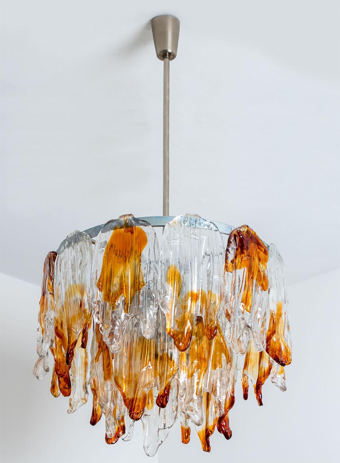 This elegant Murano glass chandelier is designed and manufactured by Mazzega, Italy in the 1960s.
The chandelier has a Chrome rod, hanging from the ceiling with orange and clear glass shades, resembling hanging leaves.
The chandelier illuminates