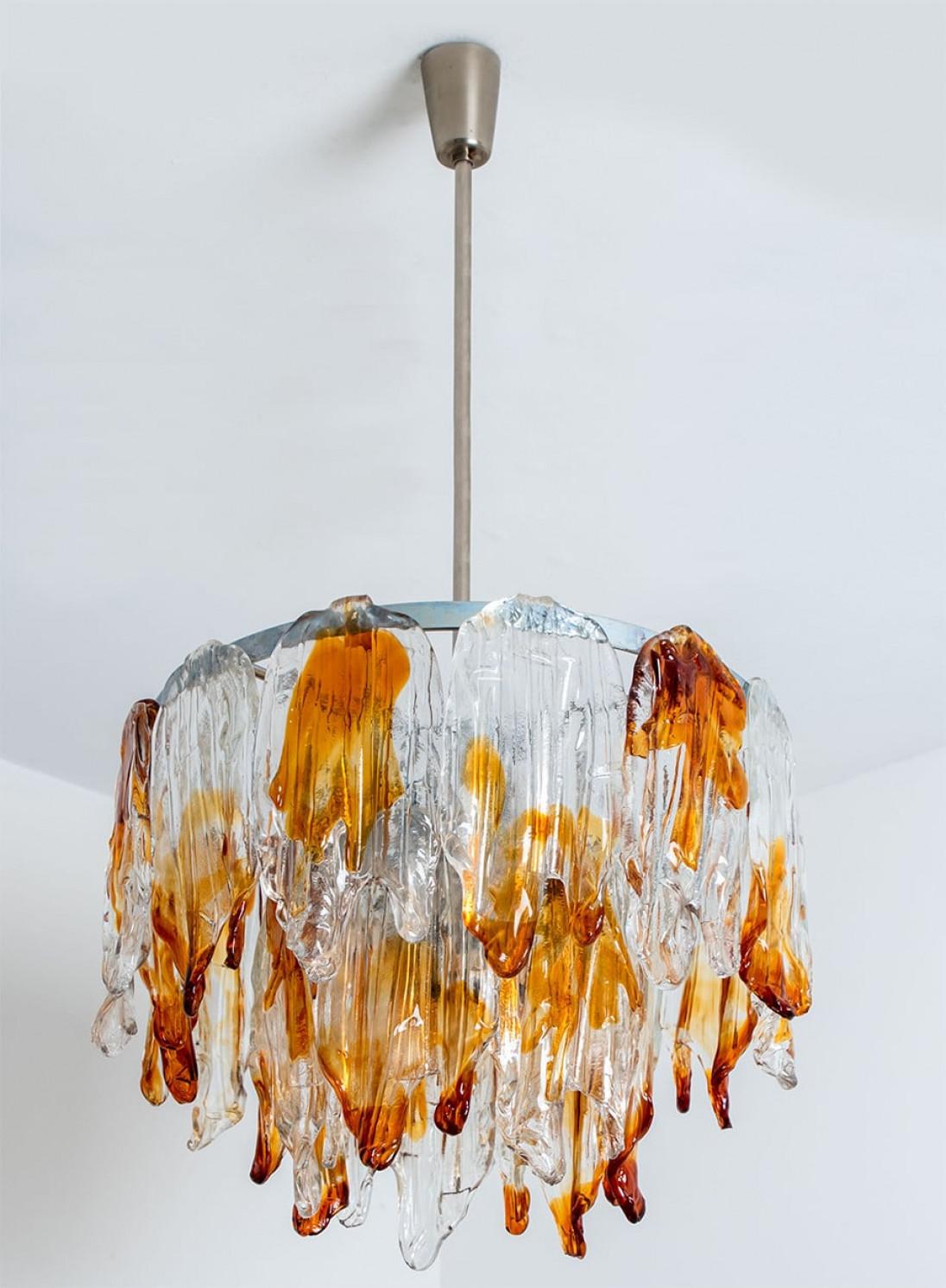 Questo elegante lampadario in vetro di Murano è stato disegnato e prodotto da Mazzega Murano, in Italia, negli anni '60.
Il lampadario ha un'asta in cromo che pende dal soffitto con paralumi in vetro arancione e trasparente, che ricordano delle