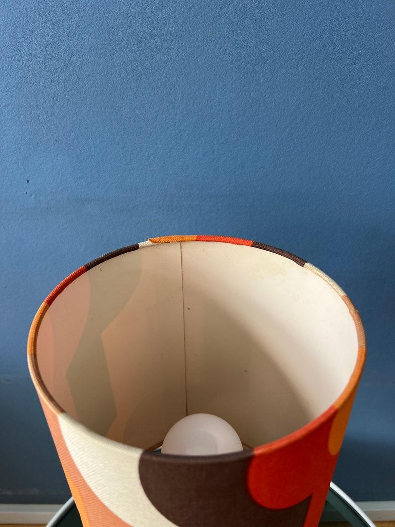 Colourful ceramic fata lava space age table lamp with orange-toned textile shade. The ceramic base has white and brown colours and a rough shape. The orange space age shade creates a funky light effect. The lamp requires one E27/26 lightbulb and