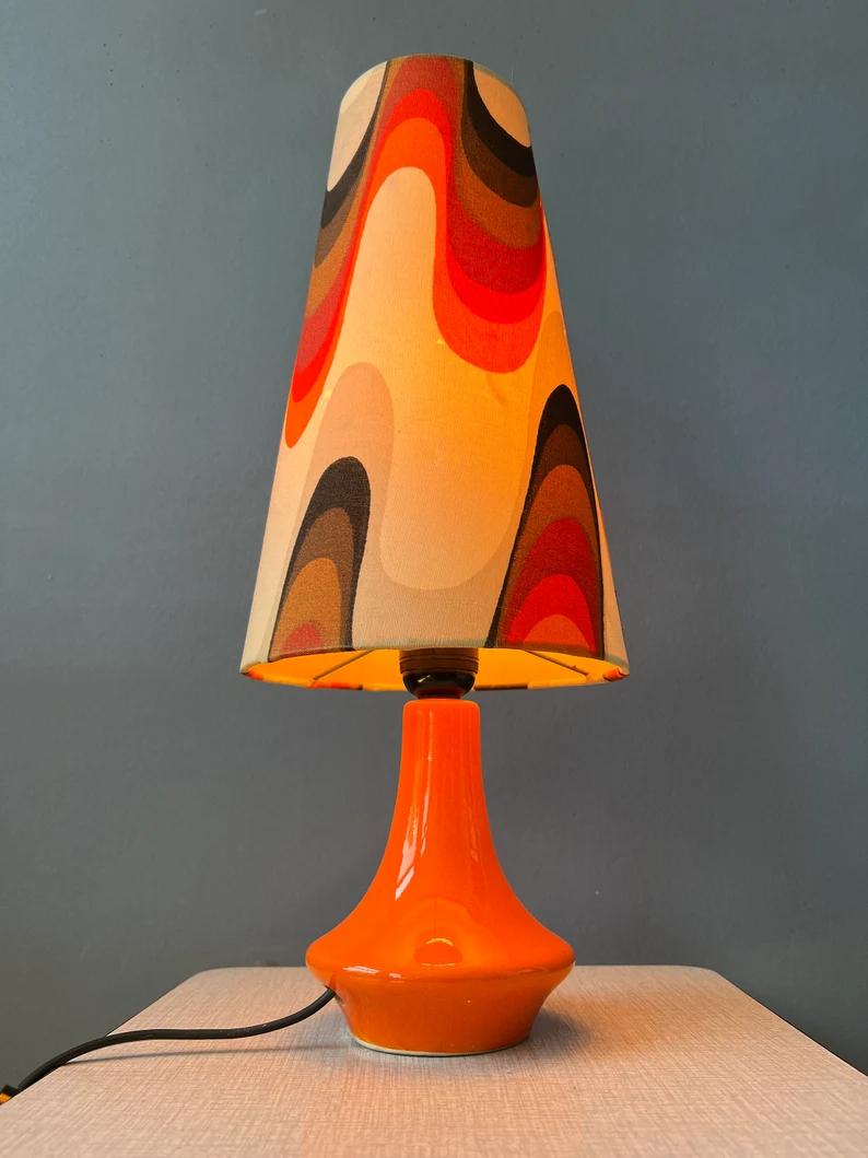 Ceramic space age table lamp with orange base and shade. The lamp requires one E27/26 lightbulb and currently has an EU-plug.

Additional information:
Materials: Ceramic, linen
Period: 1970s
Dimensions: ø Shade: 22 cm
Height: 54 cm
Condition: