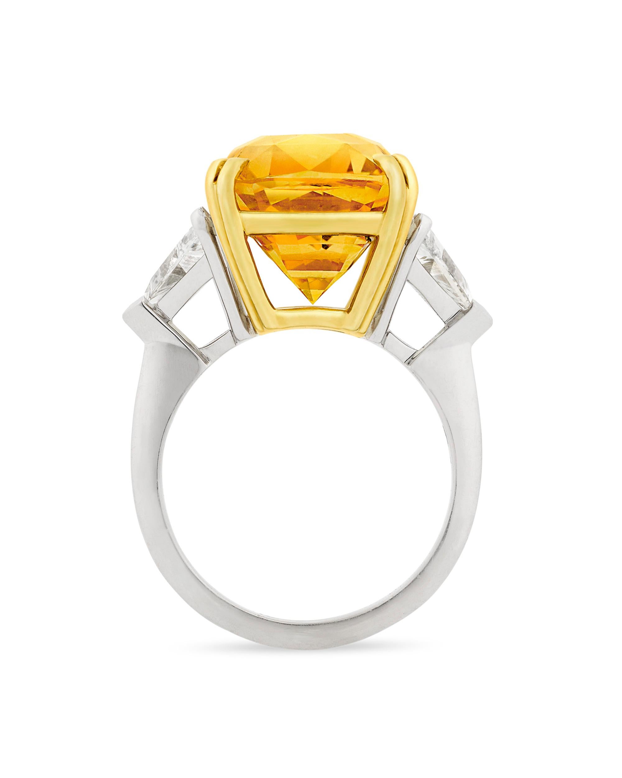 Sitting at the center of this ring is a breathtaking 15.83-carat orangy-yellow sapphire. With its stunning beauty and monumental size, this cushion-shaped gem displays amazing clarity and a highly saturated golden hue. Flanked by two white diamonds
