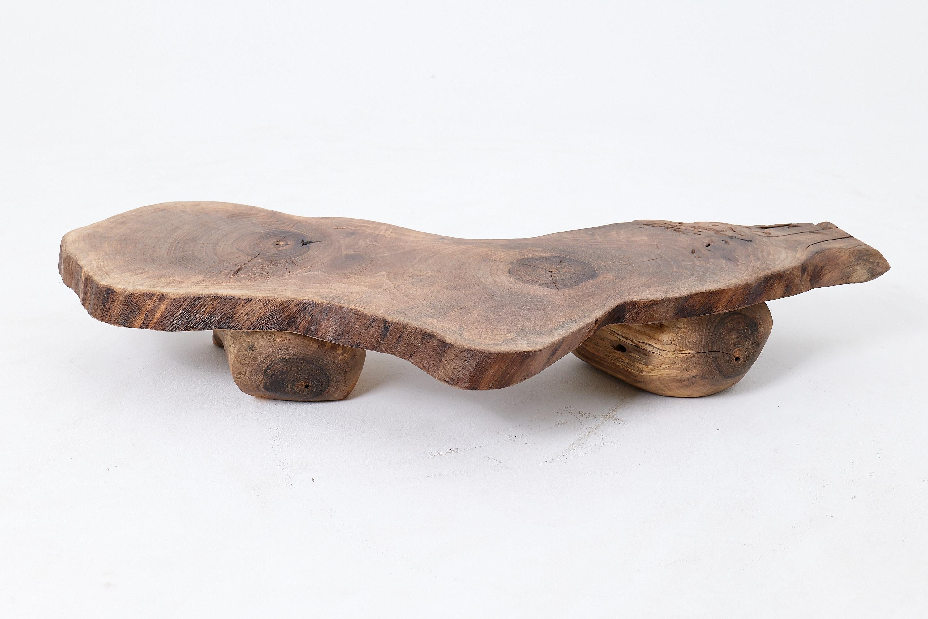 This contemplative low table is a one of a kind piece, crafted from a unique slice of solid walnut. Its form is defined by a low, grounded profile, featuring a free form slab top resting securely on two robust, natural log supports. The piece evokes