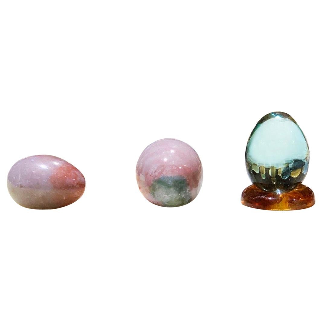 Organic modern hand carved stone and leaded blue glass egg sculptural set of 3. Gorgeous highly burnished pink granite stone eggs and luminous blue glass egg with Amber Lucite base, 4 items total in set (3 eggs + 1 base).