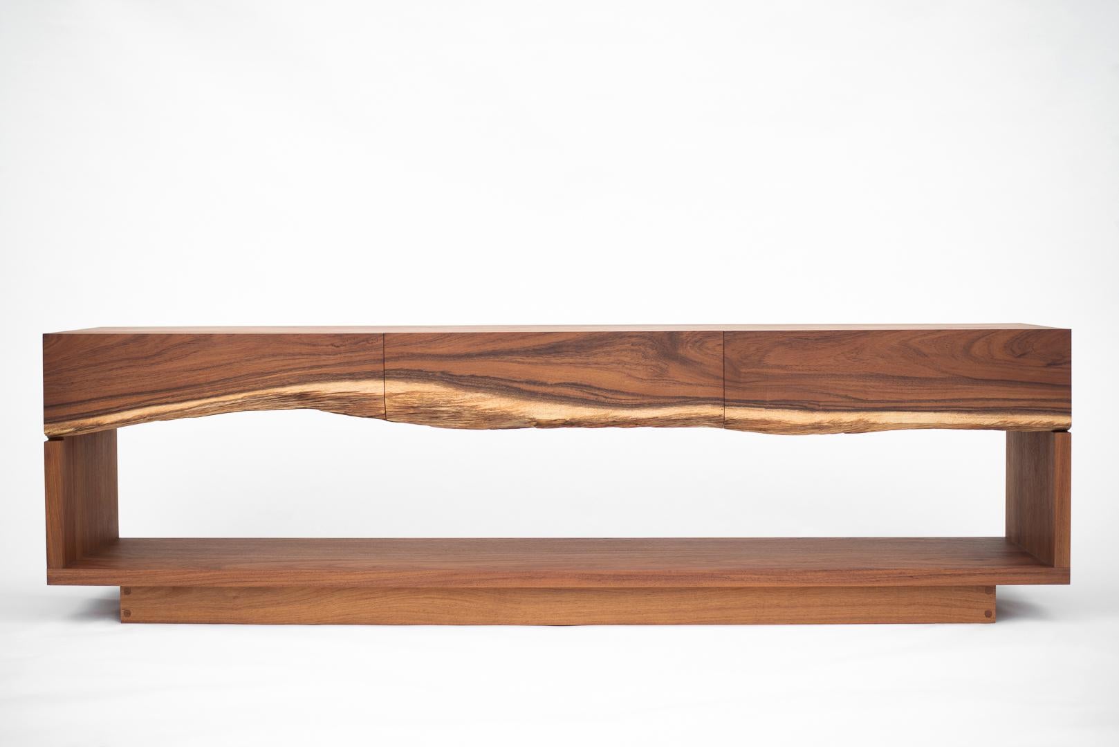 Ein Sideboard mit drei Schubladen, gefertigt aus einem einzigen Brett aus natürlichem karibischem Nussbaumholz.
Die schlichte und leichte Konstruktion dieses Möbelstücks zielt darauf ab, die Schönheit des Tropenholzes hervorzuheben und seine