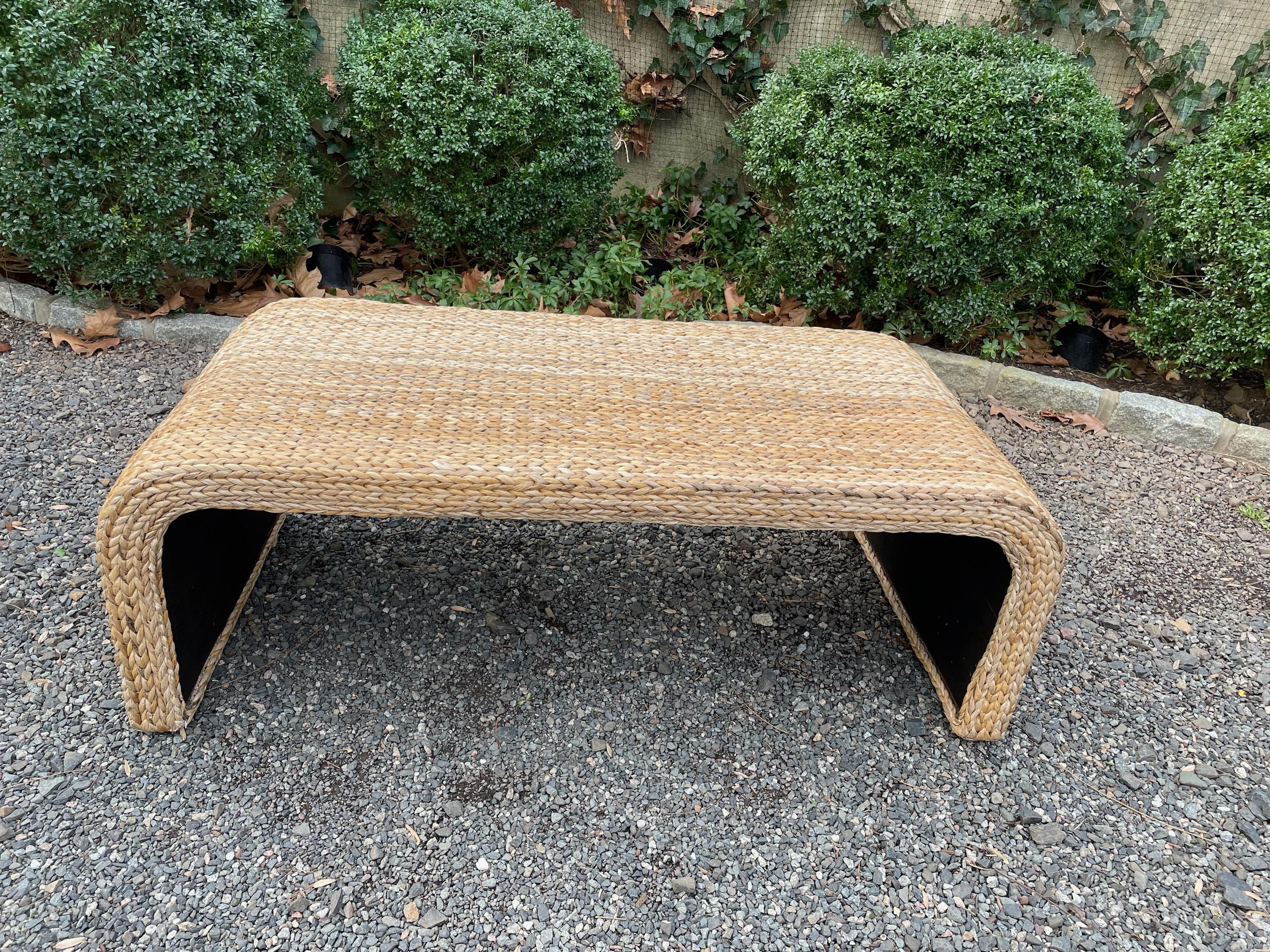 Sophisticated modernist waterfall style coffee table constructed of wood and braided natural grass. The table has arched sides and the underside is painted black.
The striations in color are natural to the grasses.