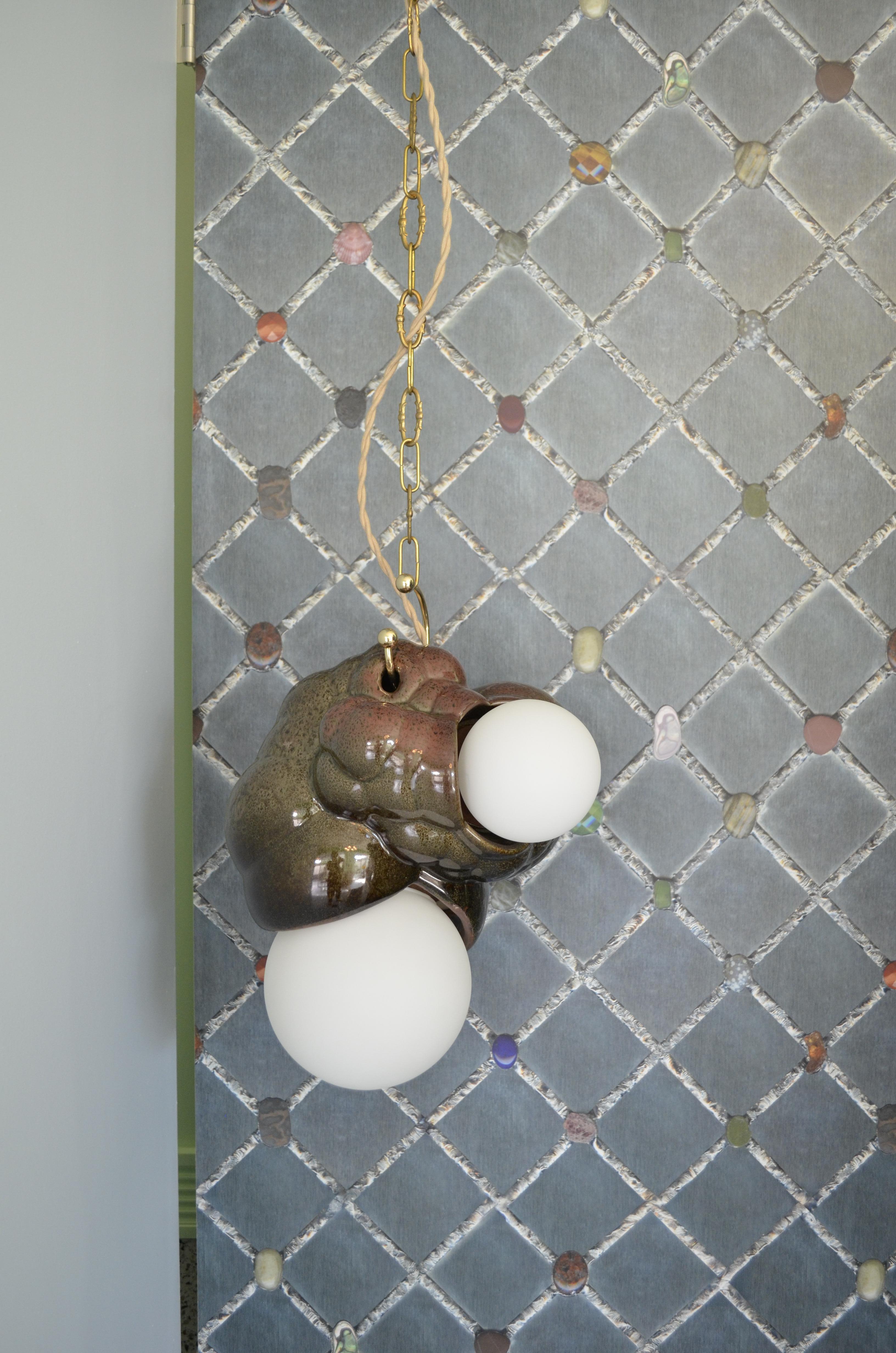 Capullo Pendant is a handcrafted ceramic pendant featuring two mouth-blown glass globes, inspired by the cocoon structures of insects as the name “Capullo” reflects. The unique hanging ring resembles a piercing, accentuating the organic and fleshy