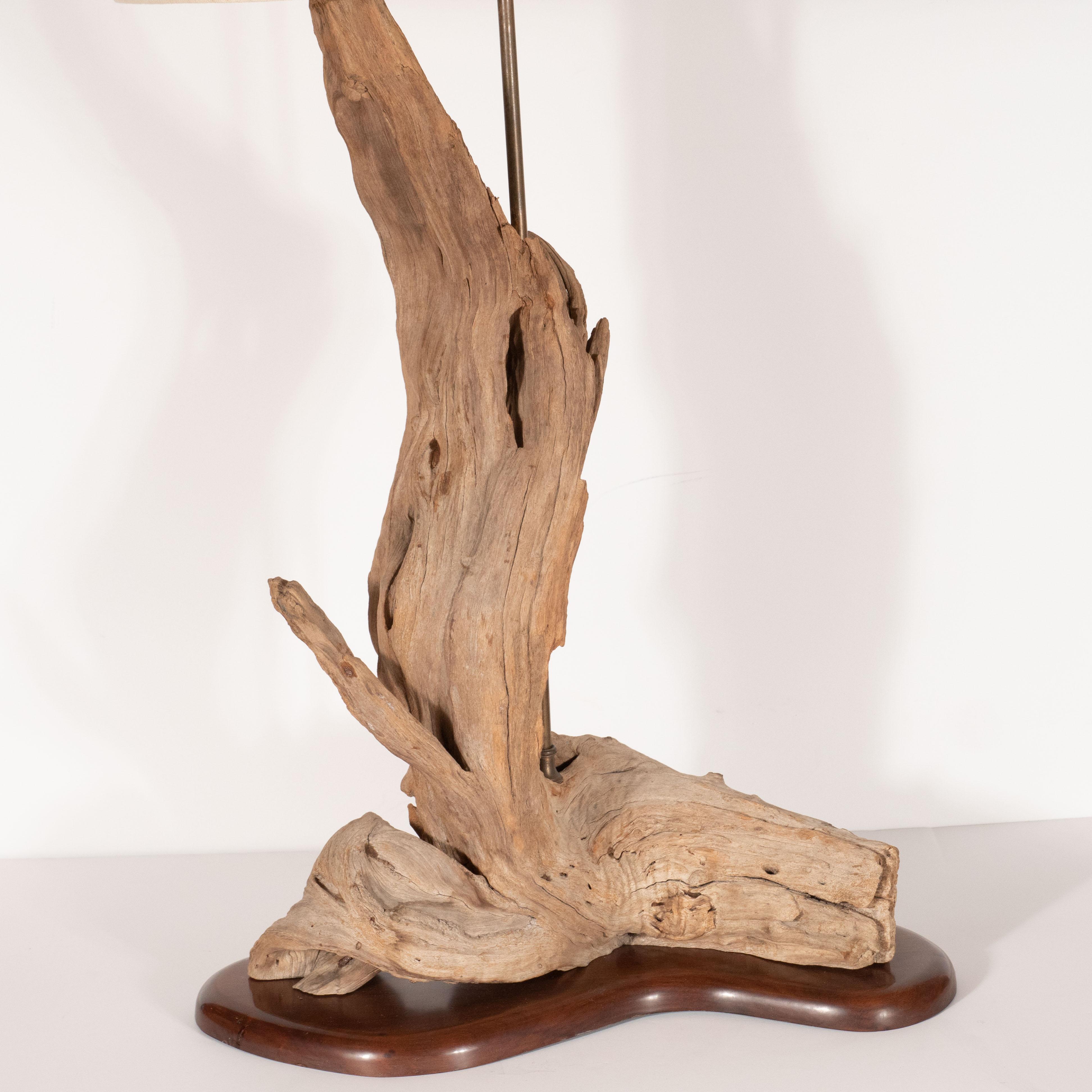 This graphic and casually elegant table lamp was realized in the United States, circa 1950. If features a sculptural body composed of unfinished driftwood resting on a amorphic stylized 