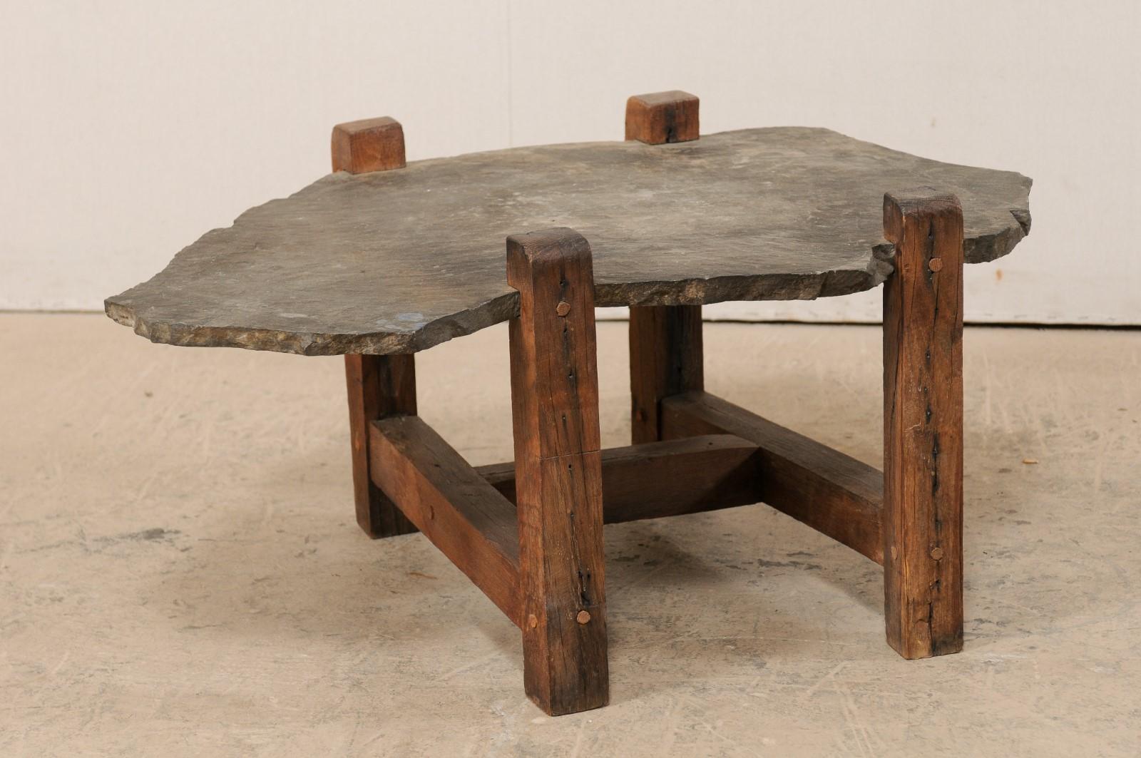 A vintage American wooden coffee table with slate top. This rustic coffee table has been designed with four squared legs, set at each corner, having notched cut-outs just below their tops, where a slab of natural slate stone makes up the table top.