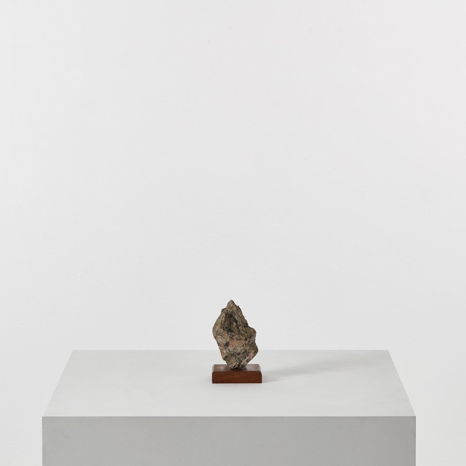 A sculpture in the loosest sense, this weighty stone fragment is knitted with geologic seams. The rough surface has a rich minerality in contrast with the chamfered wooden plinth. There is some glue visible where the stone and wood connect. 

