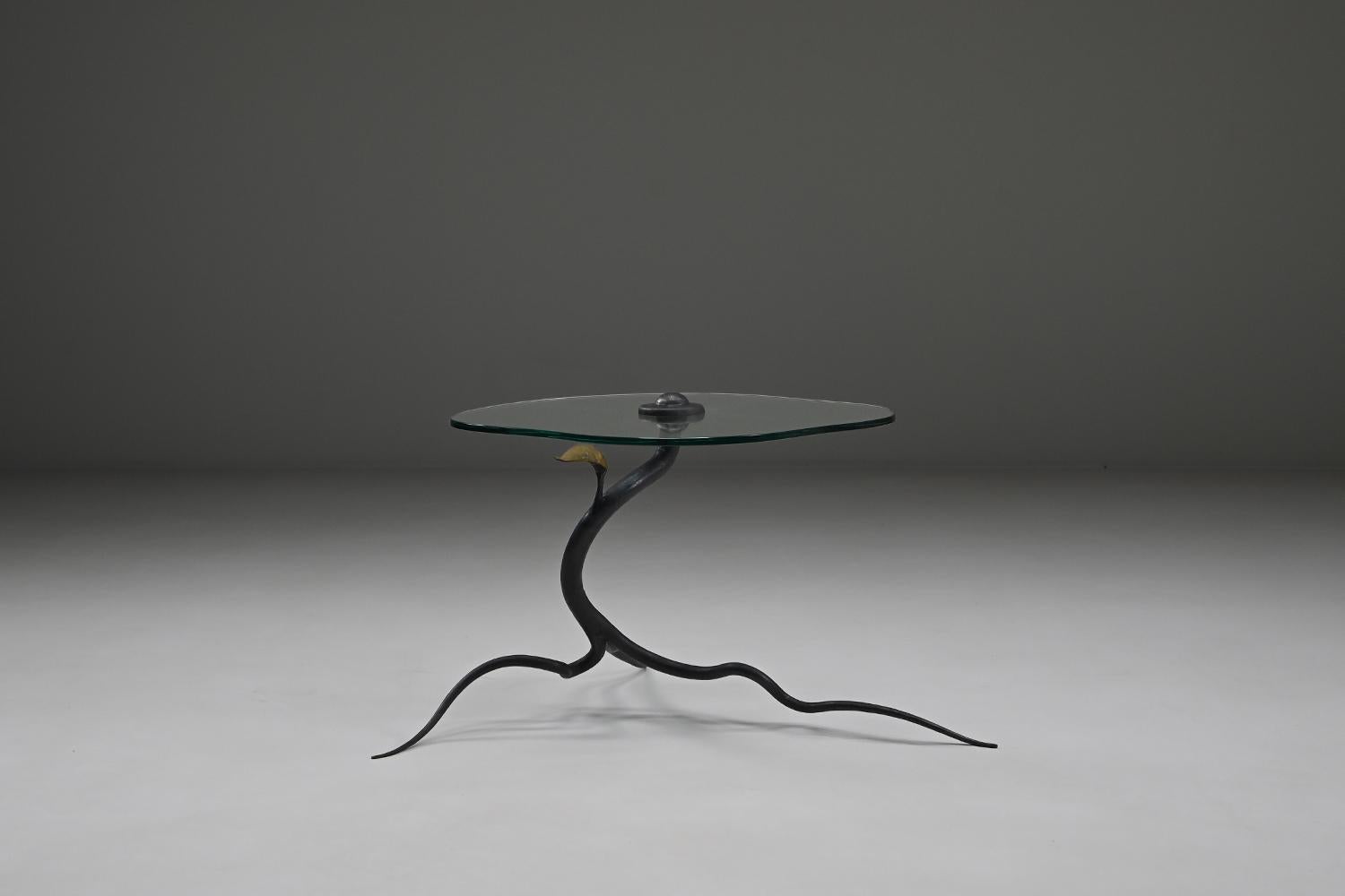 Organic wrought iron side table, 1970s Belgium. This side table features a hand-forged wrought iron base shaped like a branching root that extends in three directions, supporting a clear organic shaped glass tabletop. A single gold-colored leaf