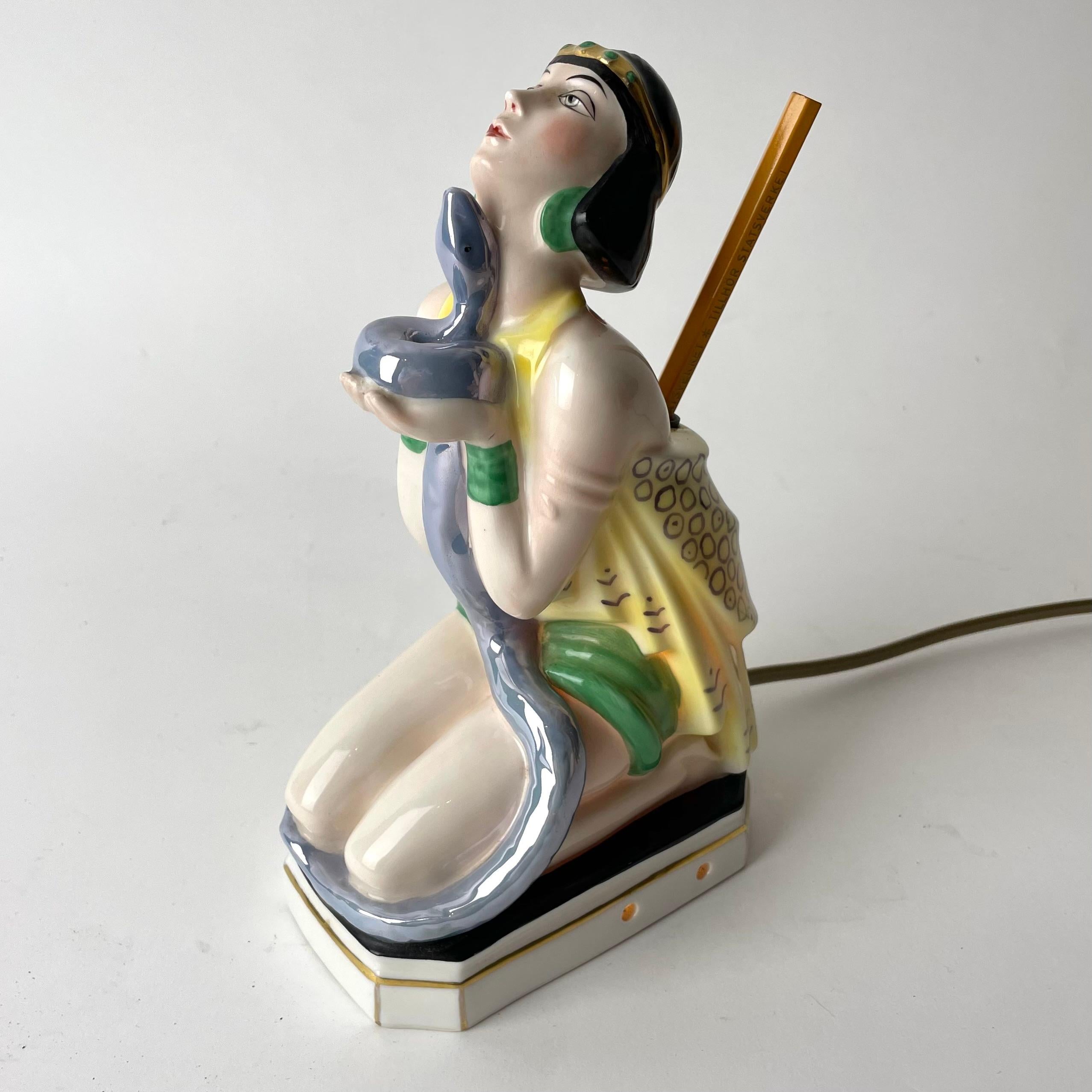 Art Deco Table Lamp with Pen Stand, depicting lady with oriental dress facing upwards while holding a snake. 
A charming table lamp from the 1920s. The woman kneels upon a black and white ceramic base with golden details. A blue snake stretches from