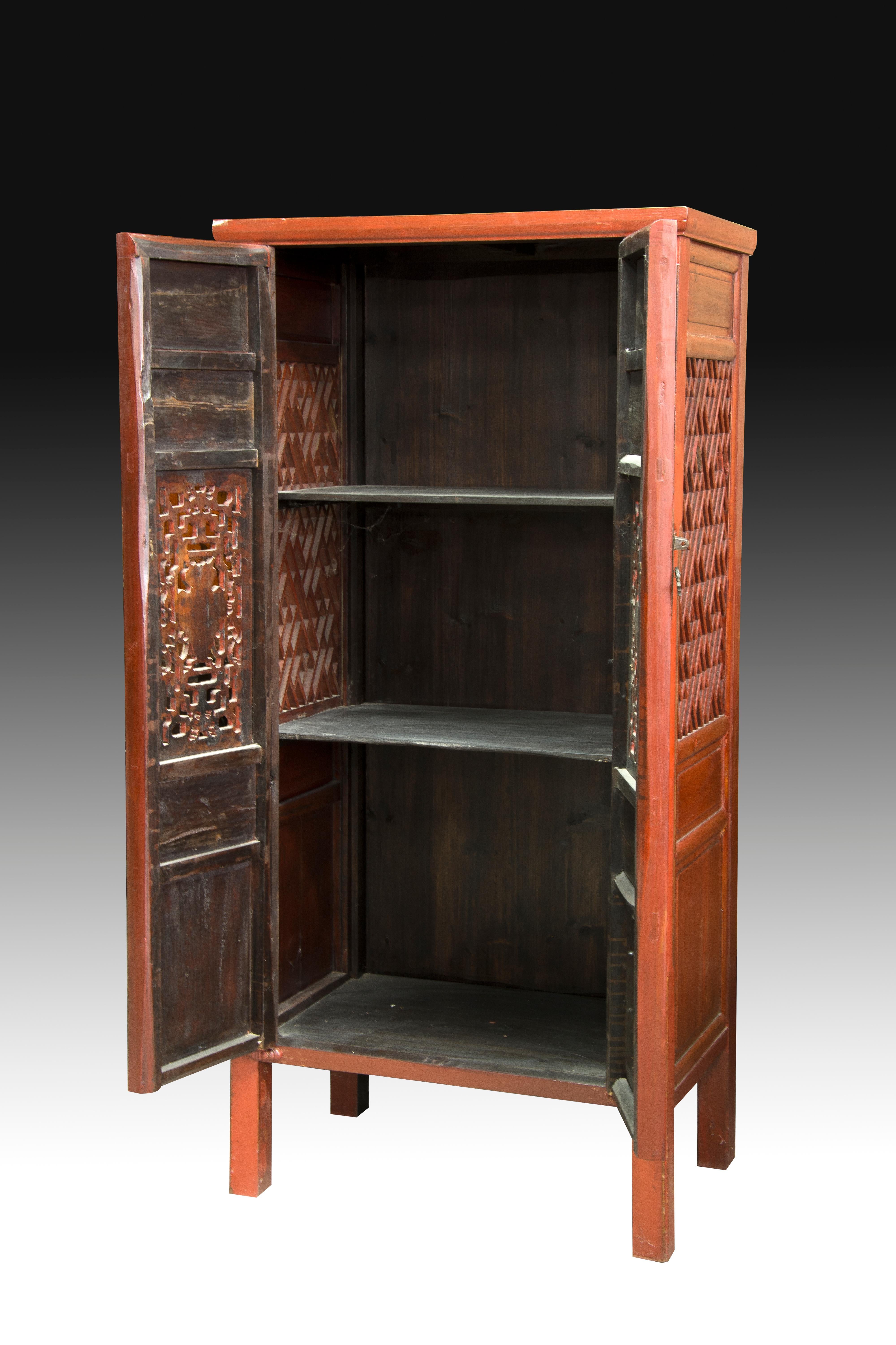 Tall carved wooden wardrobe decorated on the sides with simple geometric shapes enhancing a geometric fretwork composition and, on the front, with various rectangular spaces with scenes, geometric elements set around figurative scenes and floral