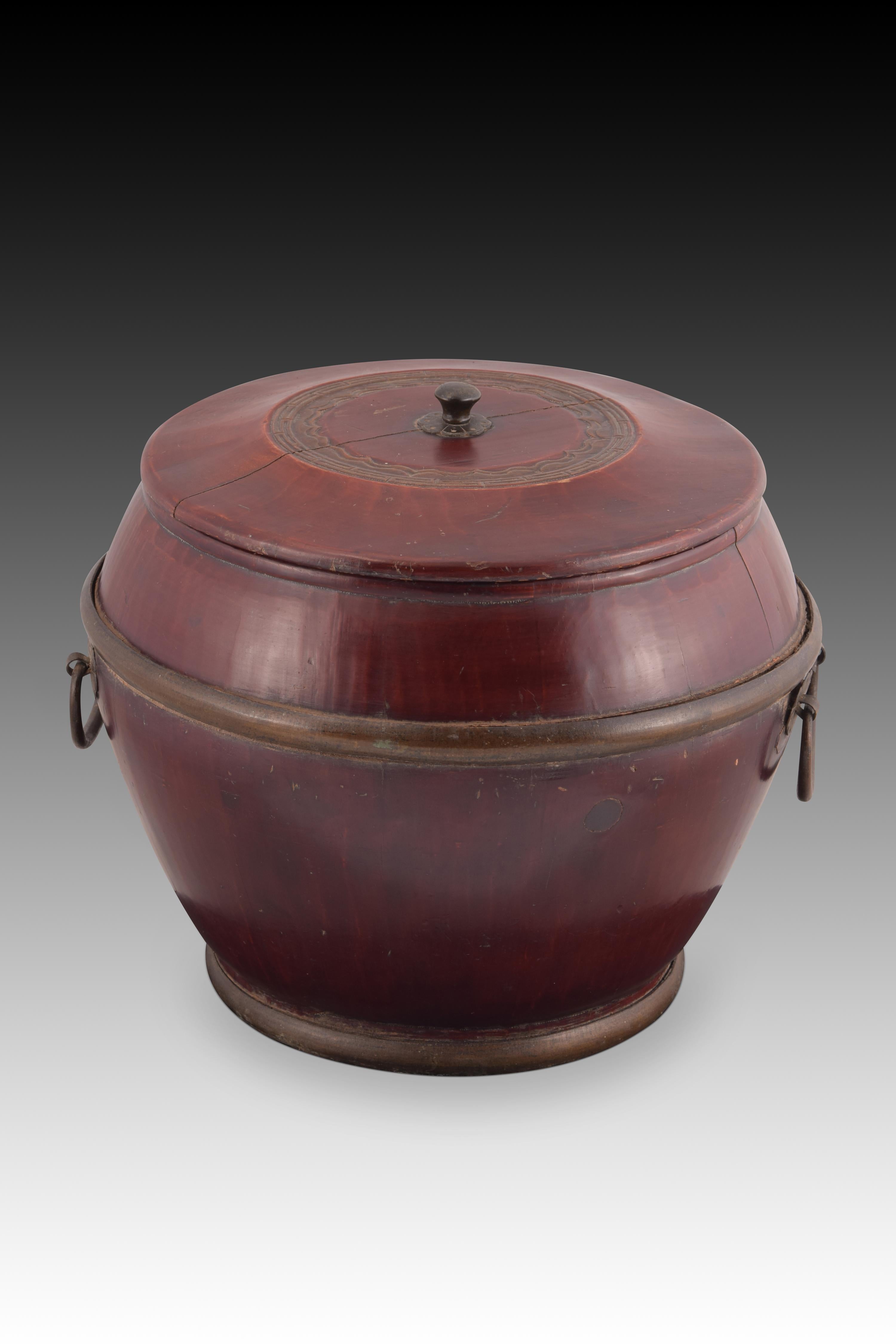 Oriental container for rice. Wood and metal. China, towards the beginning of the 20th century. 
Chinese container for storing rice, made of carved and varnished reddish wood, with a flattened ovoid body and a flat lid, slightly raised towards the