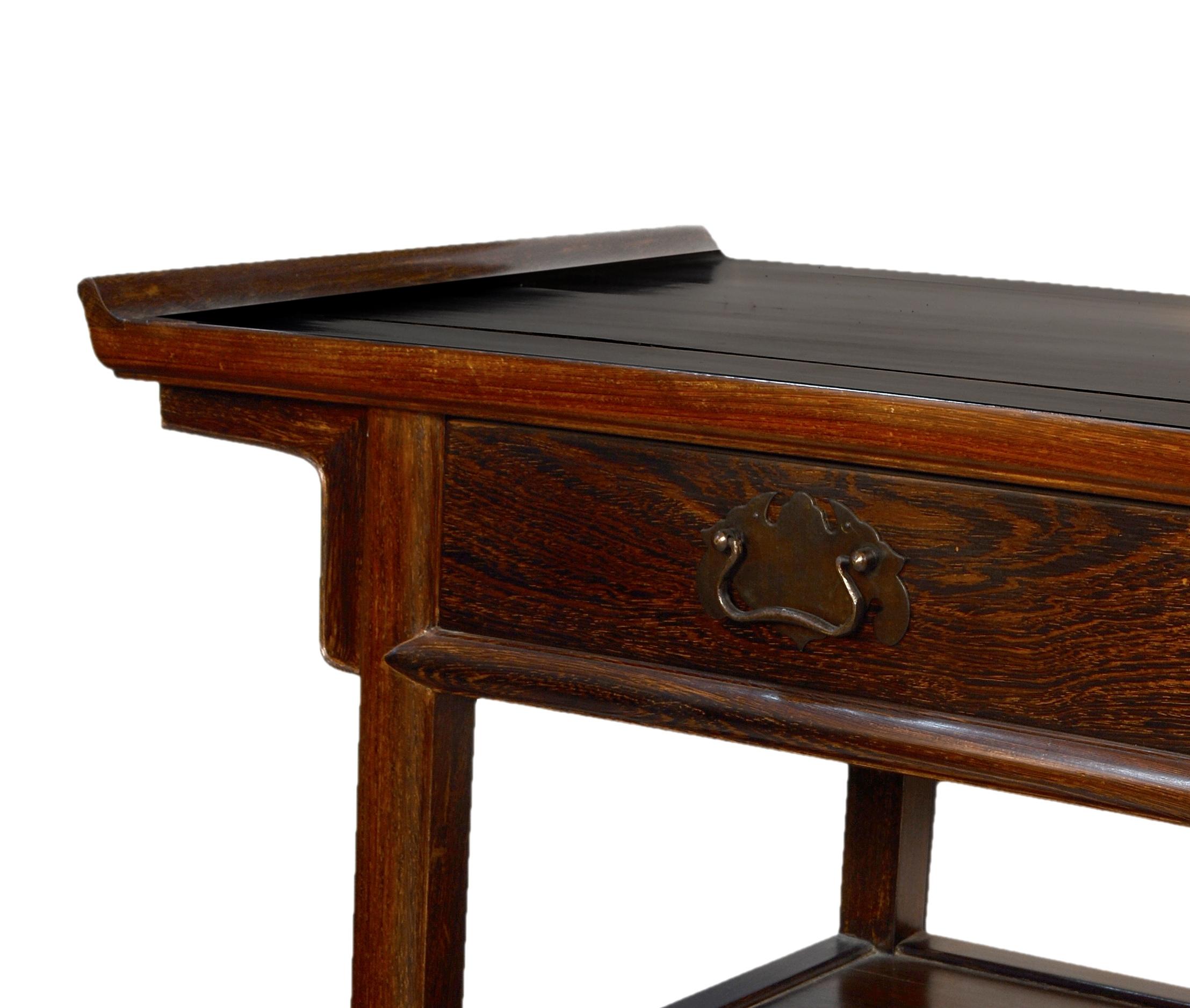 Rectangular board table decorated with some elevations on the sides that has a board-shaped chambrana on the bottom and a drawer to the front with metal hardware. An influence of Chinese furniture can be seen in the arrangement of legs, drawer and