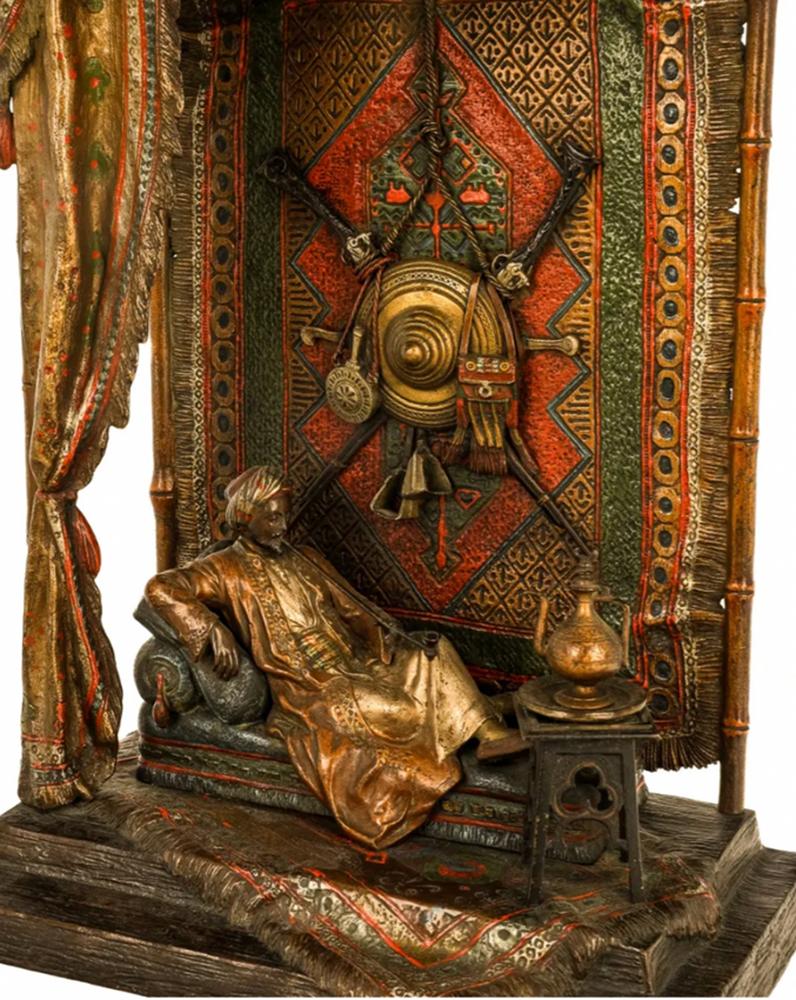 A Sultan reclining on a chaise within an open Bedouin tent made of draped rugs and a bamboo frame. A shield, swords and ornate bags, hang from the intricately woven rug above him. He smokes a simple pipe, wearing golden robes and a matching turban.