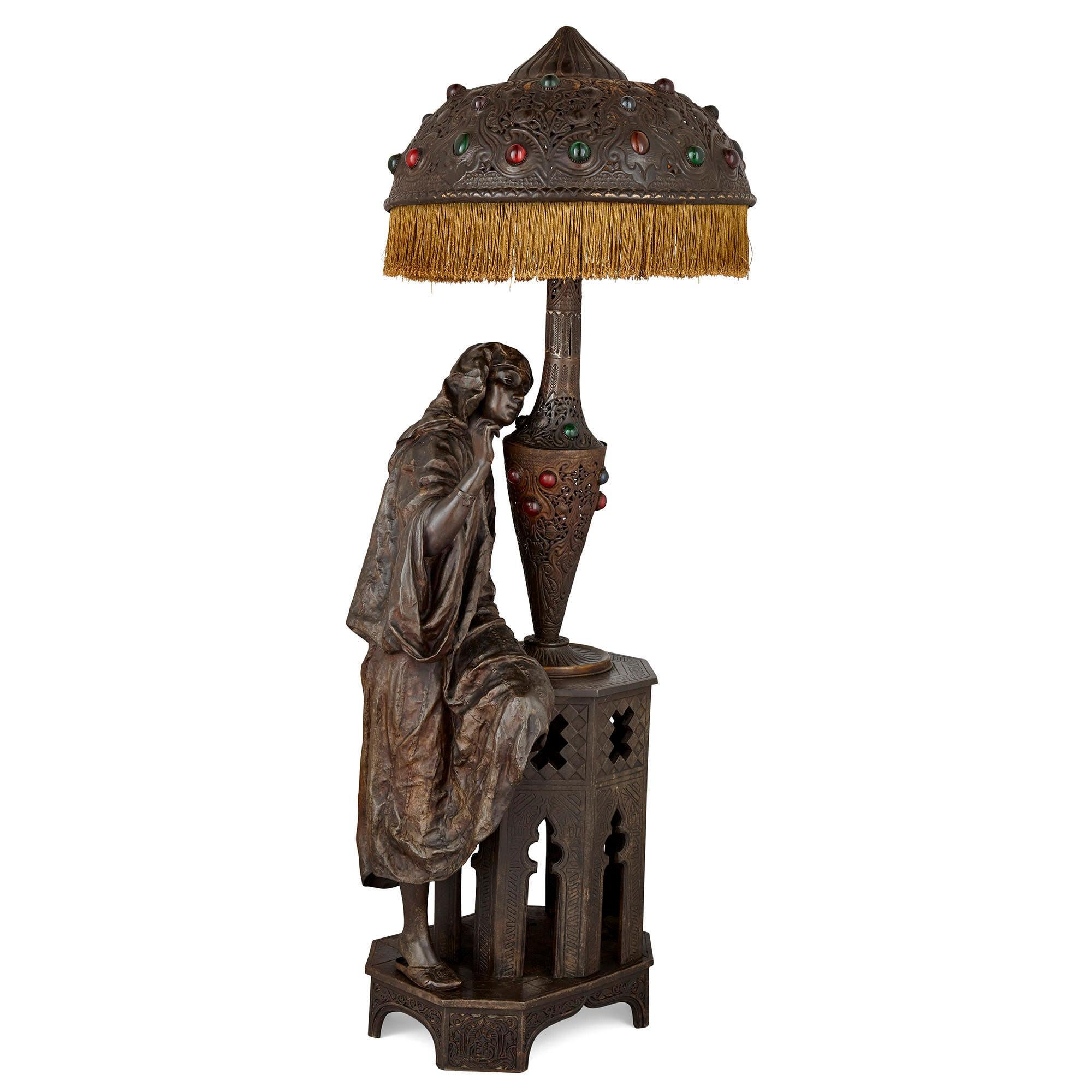 This Belle Époque period orientalist lamp features a charming ensemble of elements. Most strikingly, a female figure in orientalising dress leans against a Moorish side table, upon which sits a table lamp surmounted by a shade and ornamented with