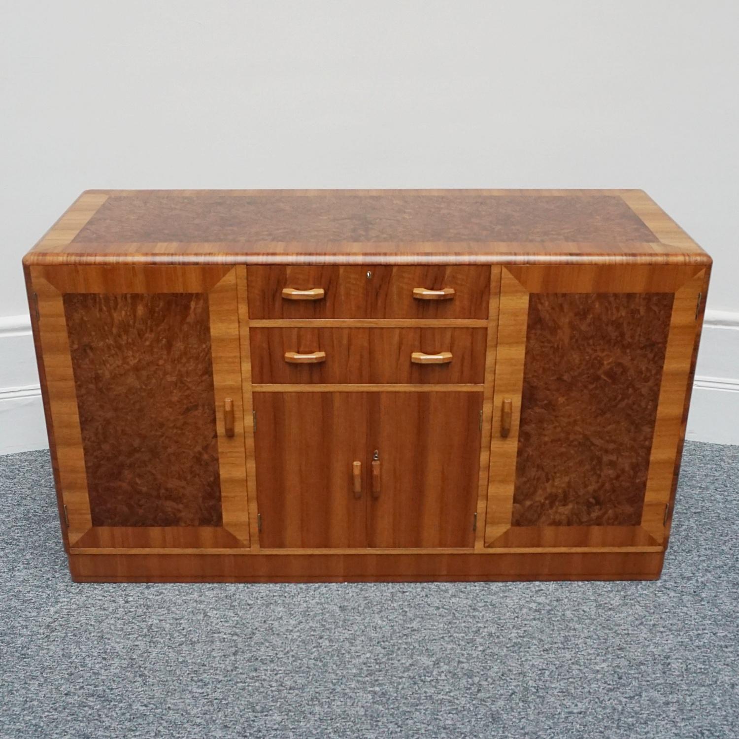 An Art Deco sideboard by Heal's of London. Burr walnut veneered with figured walnut banding on solid mahogany/ Original walnut handles. Two central upper drawers with lower cupboard section. To either side cabinet doors open to glass shelved