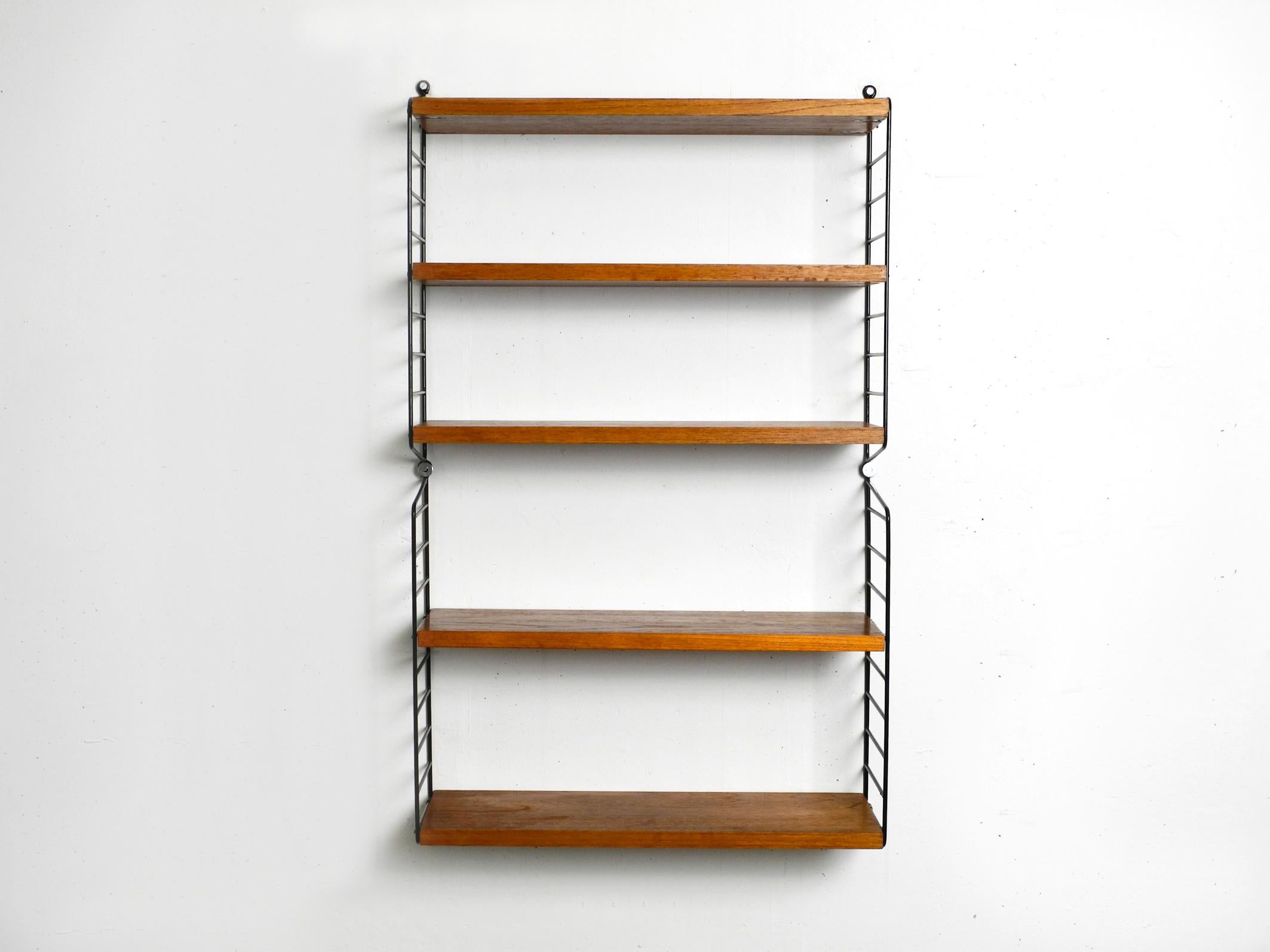 Original 1960s Nisse Strinning wall hanging shelf with five solid wood shelves with teak veneer.
This string shelf has 20cm narrow shelves with a width of 58 cm.
4 metal ladders with black plastic coating.
All four ladders are in good condition. No