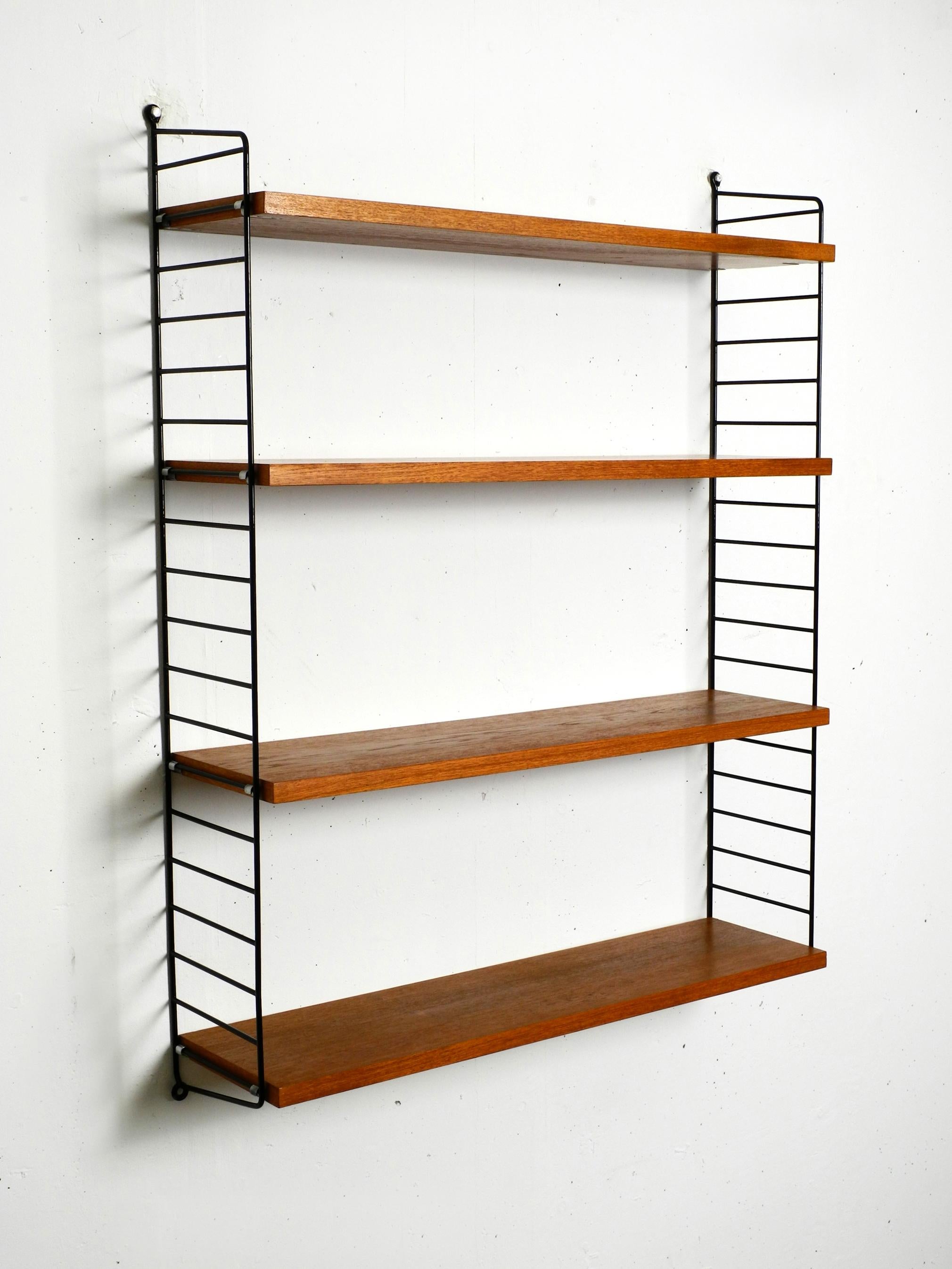 Original 1960s Nisse Strinning wall hanging shelf with four solid wood shelves with teak veneer. 2 metal ladders with black plastic coating. All four ladders are in good condition. No rust. The plastic coating is still perfect.
With slight signs of