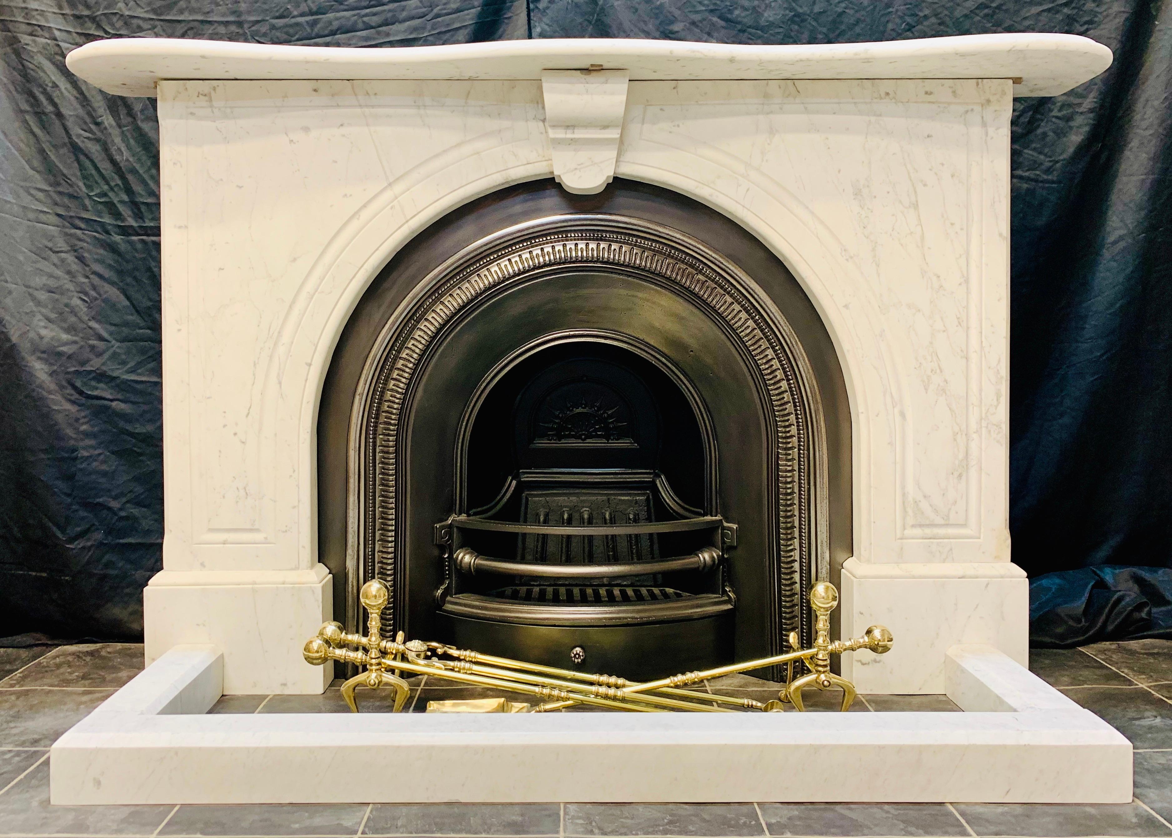 A charming and versatile 19th century Victorian lightly veined pencil Carrara marble arched fireplace surround. A serpentine shaped shelf sits above a pair of carved spandrel arches centred by a well carved keystone corbel, all resting on moulded